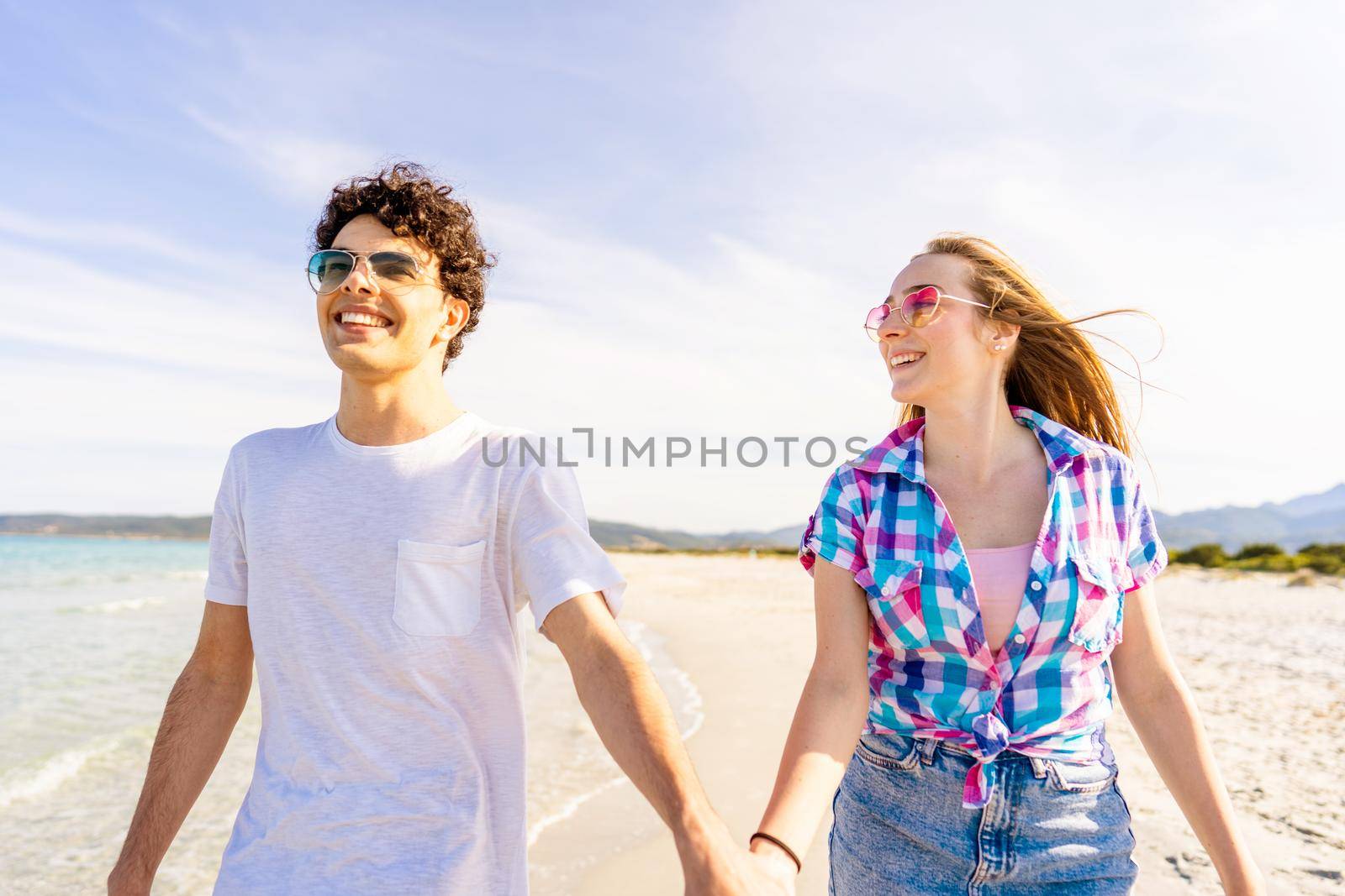 Carefree youth concept: young very happy gen z real couple holding hands tenderly walking on the seashore of tropical beach in summer with sunglasses. Girl in love looks intently at her boyfriend