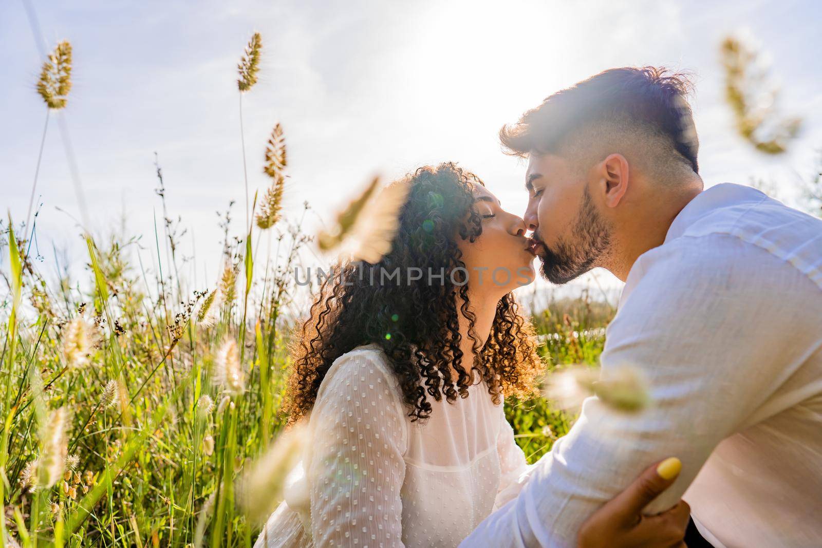 Romance scene of mixed-race couple in love kissing in backlight effect among flowers and high grass with flare and reflection sun effect at sunset or sunrise. Bearded macho young man flirting