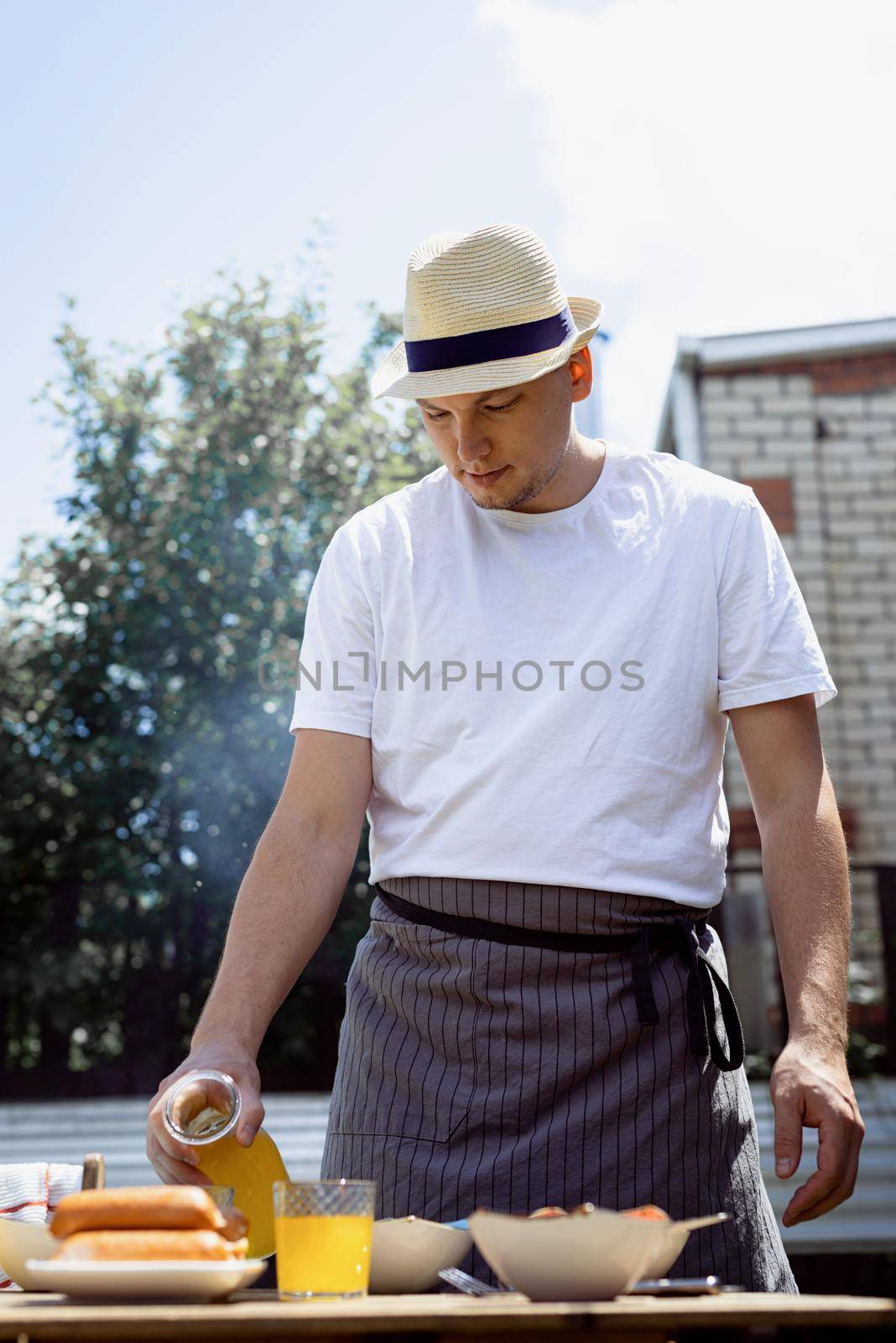 Young man preaparing table for picnic , holding bottle with lemonade in sunny summer afternoon. Backyard barbecue