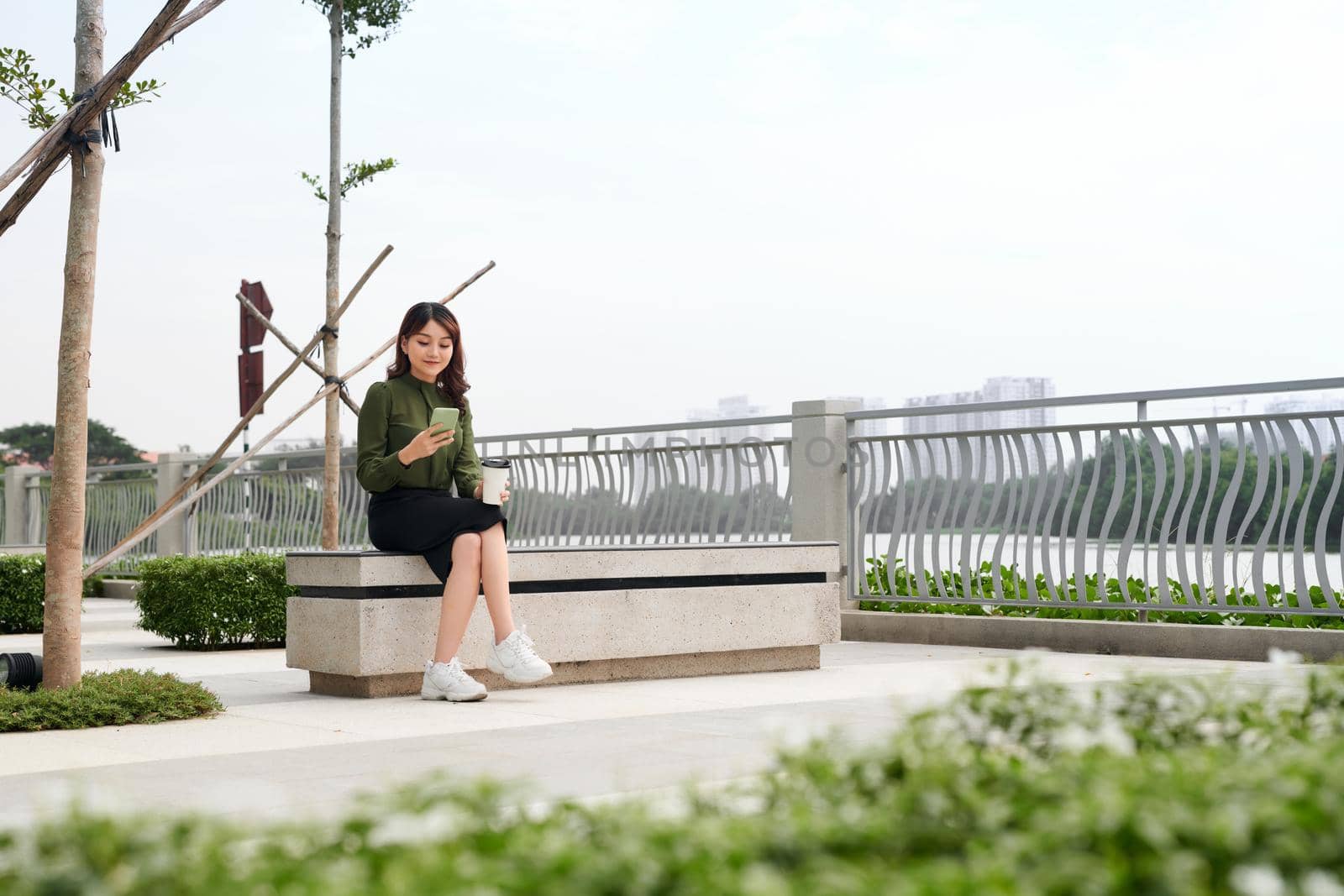 Happy young woman with a disposable coffee cup sitting on the park bench and using her smartphone
