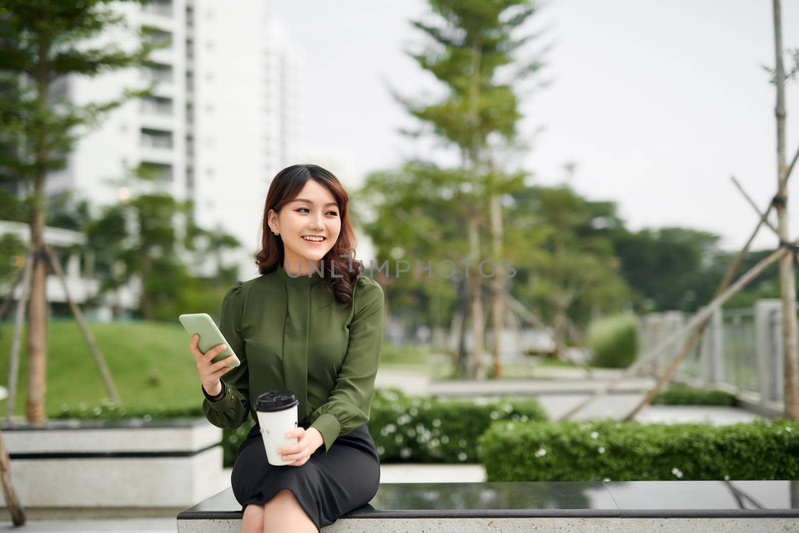 Beautiful business woman in casual green outfit sitting in park, holding coffee and talking on a phone. Daylight, outdoors.