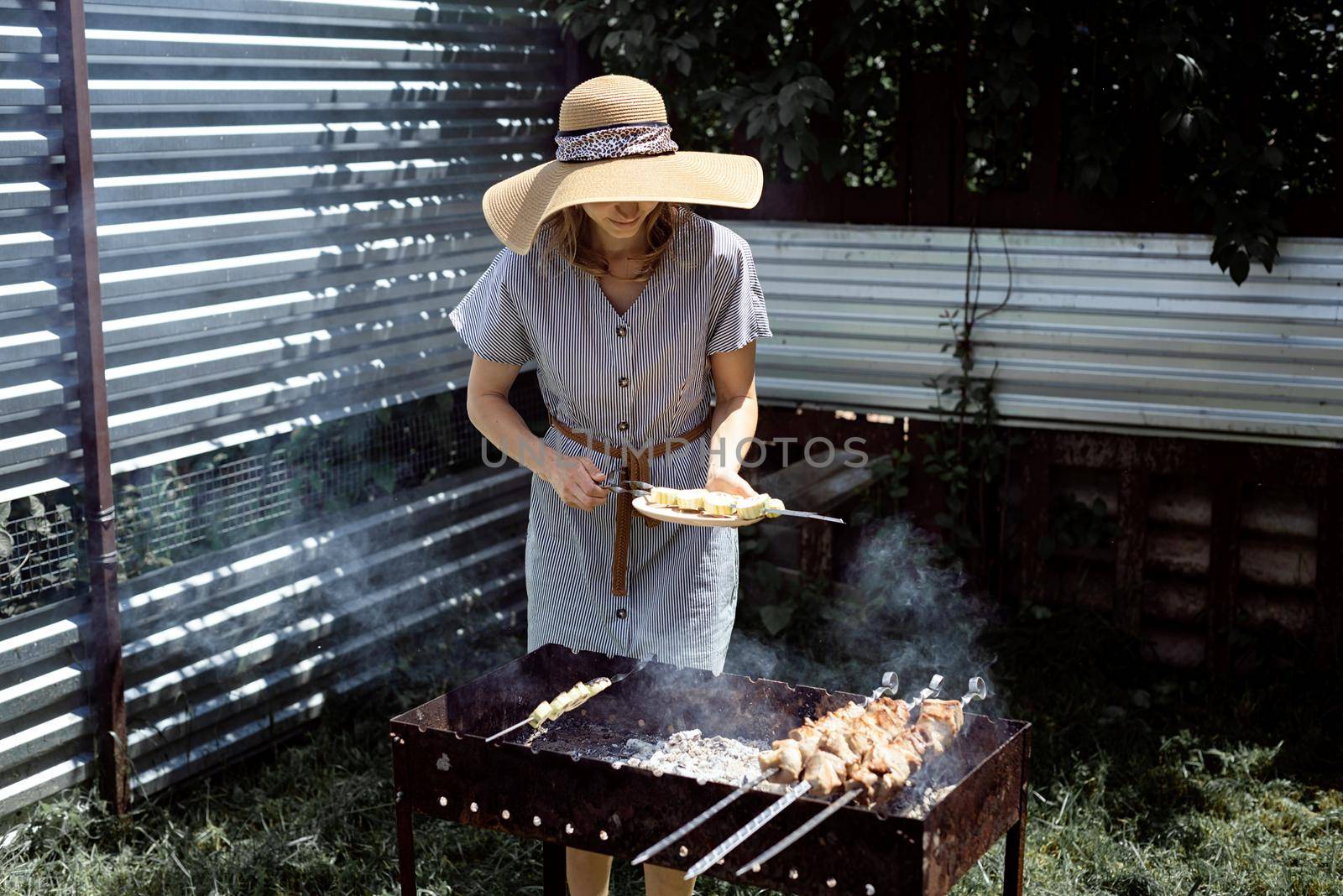 Young woman in summer hat grilling meat outdoors in the backyard by Desperada