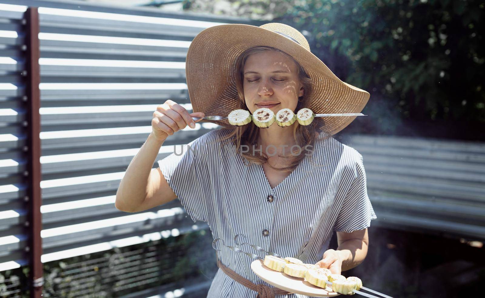 Young woman in summer hat and dress grilling meat and vegetables outdoors in the backyard. Young woman in summer hat smelling grilled vegetebles outdoors