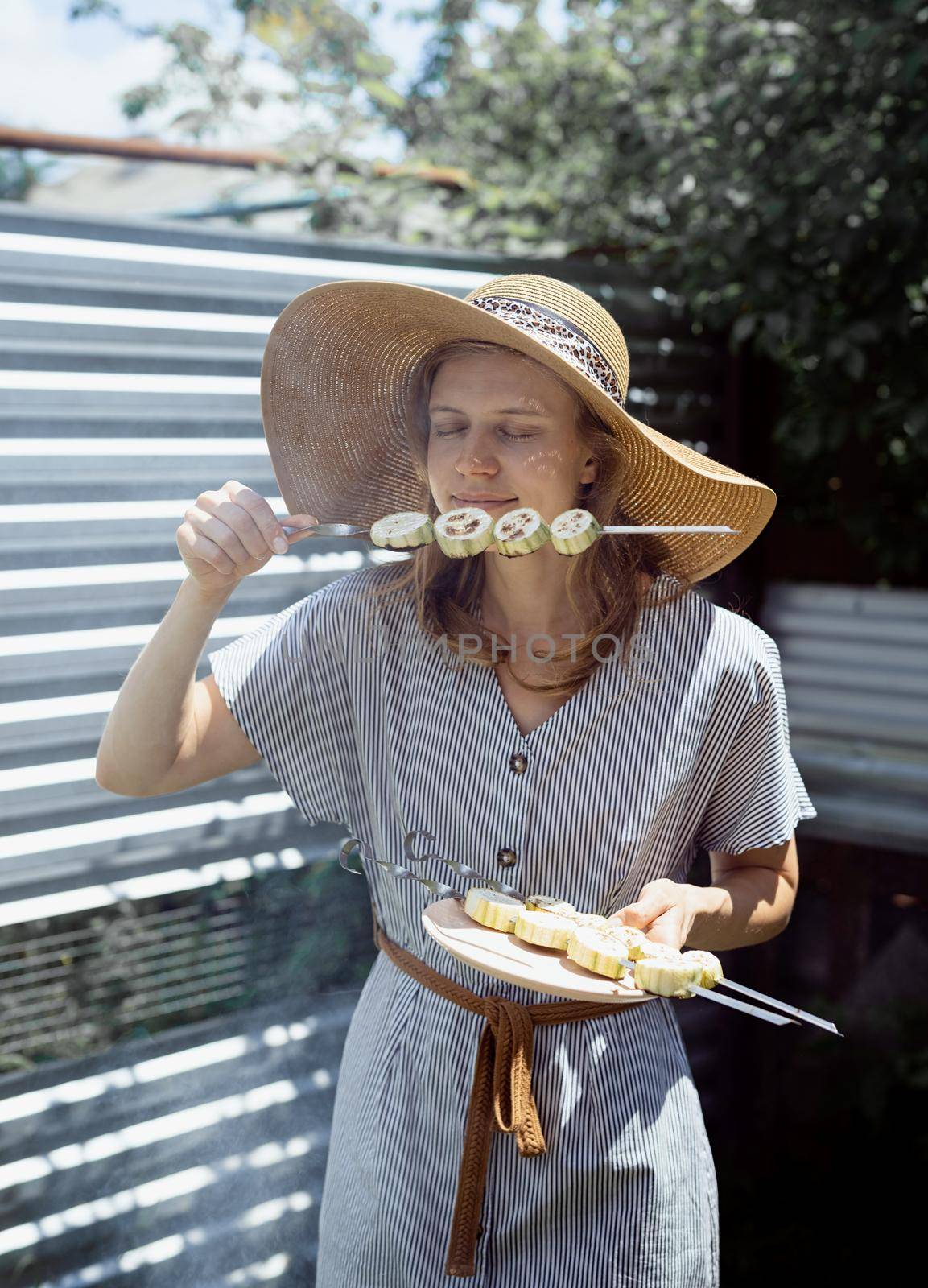 Young woman in summer hat smelling grilled vegetebles outdoors by Desperada