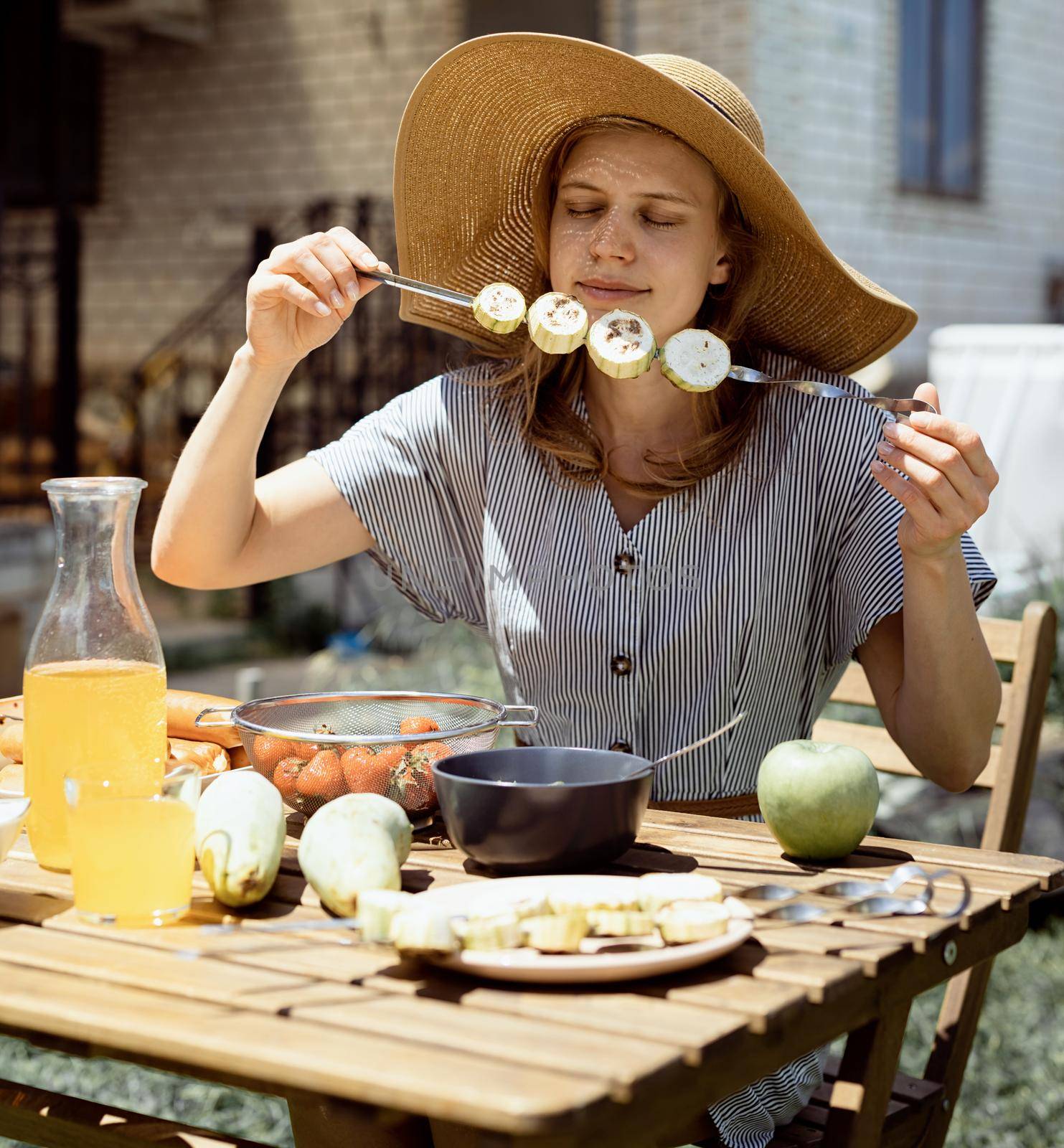 Young woman in summer hat sitting at the table, eating grilled vegetebles outdoors by Desperada