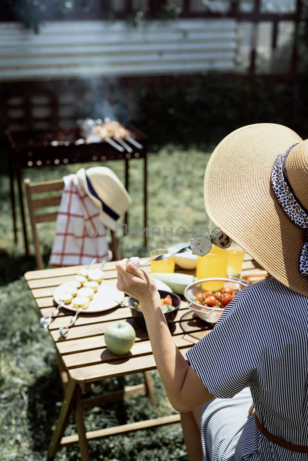 Young woman in summer hat sitting at the table, eating grilled vegetebles outdoors by Desperada