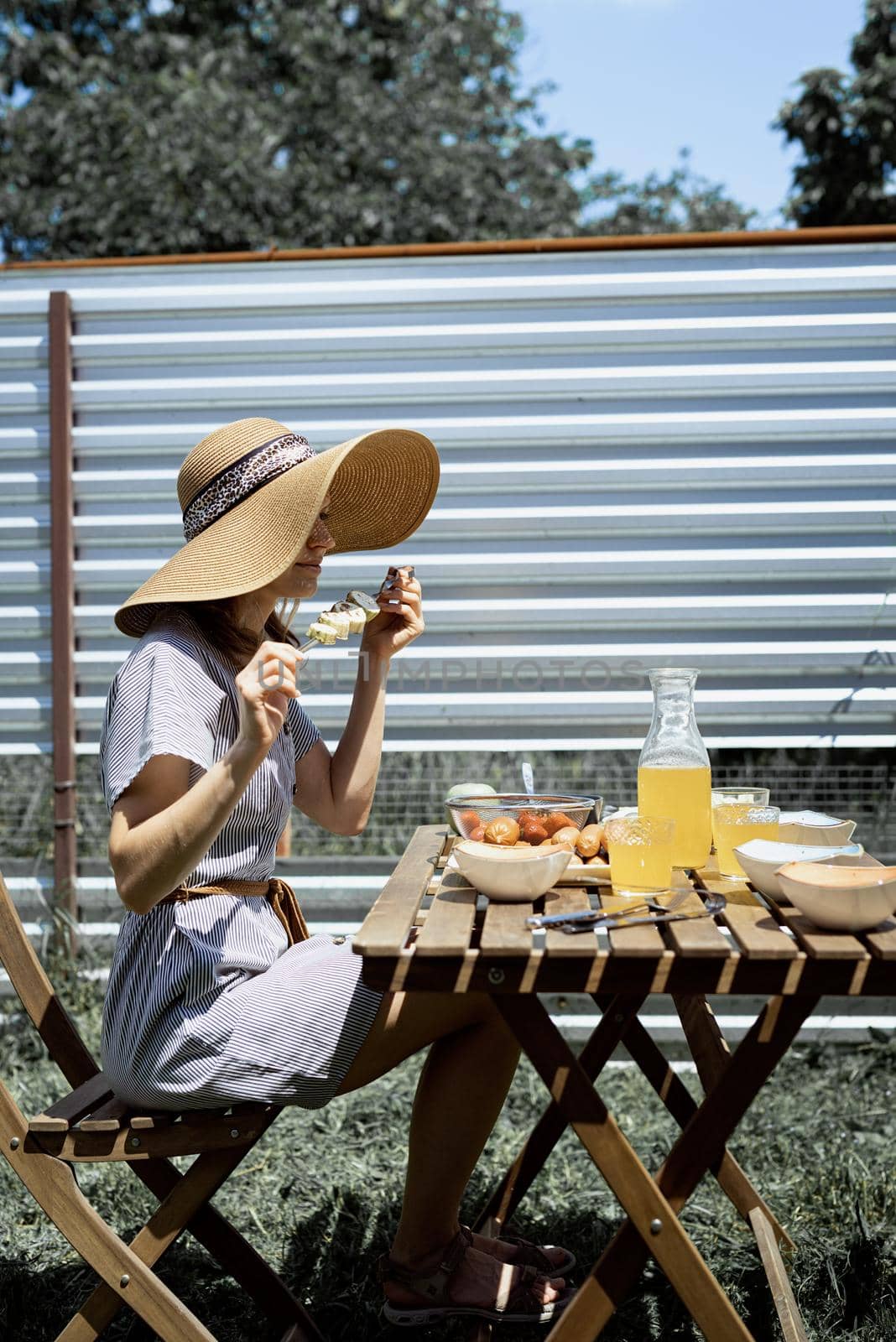 Backyard barbecue. Young woman in summer hat sitting at the table, eating grilled vegetebles outdoors