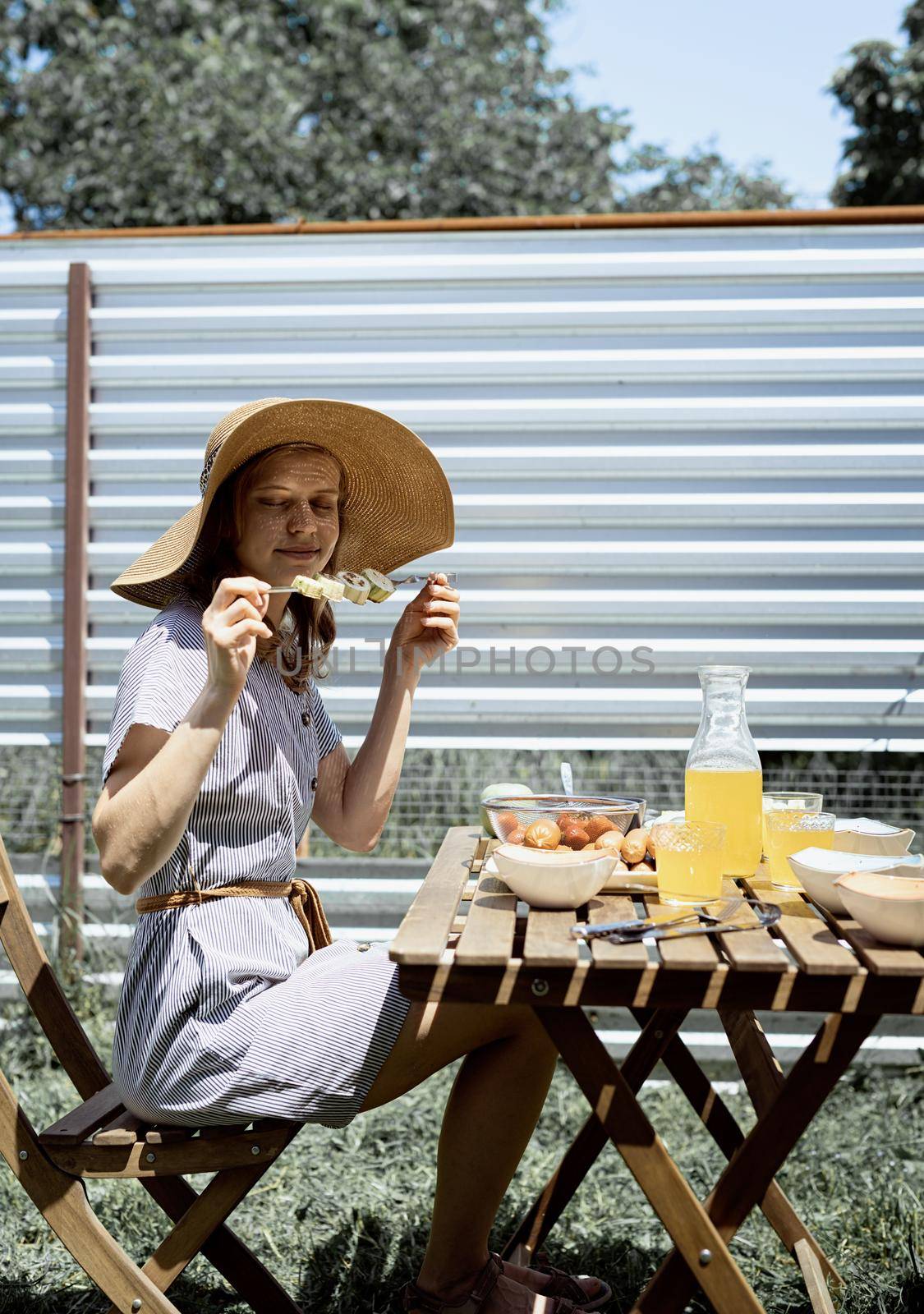 Young woman in summer hat sitting at the table, eating grilled vegetebles outdoors by Desperada