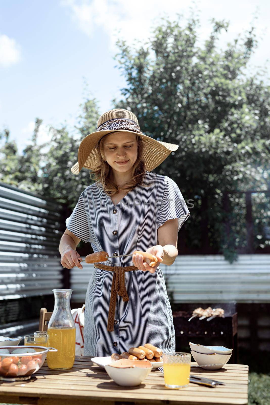 Young woman in summer hat and dress grilling meat and vegetables outdoors in the backyard.