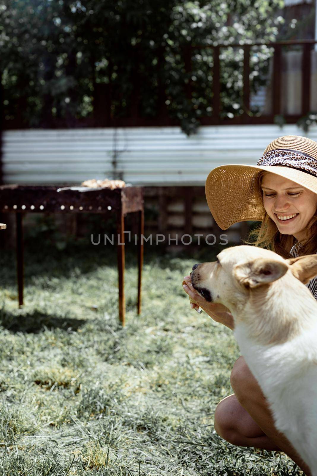 Young woman in summer hat grilling meat outdoors in the backyard, sitting with her dog by Desperada