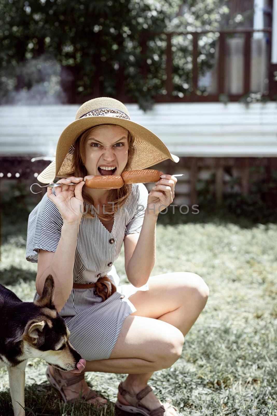 Young funny woman in summer hat grilling meat outdoors in the backyard, sitting with her dog by Desperada