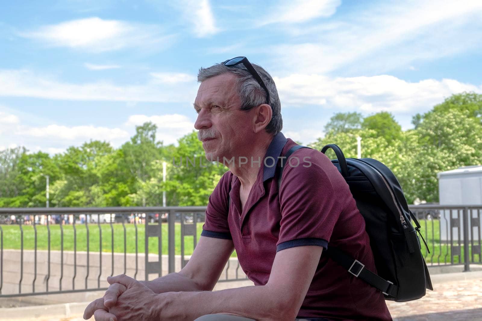 Close-up of an adult retired man in summer clothes and with a backpack sits and rests against the background of a summer cityscape. Life style. Selective focus.