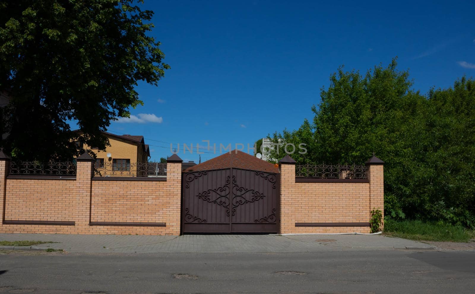 Light brick fence with iron wrought iron gates on the street against a background of wood and blue sky by lapushka62