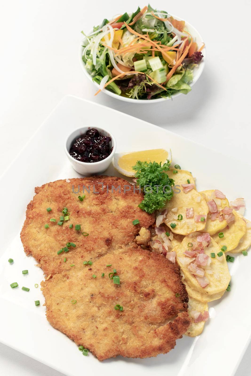 german breaded organic pork schnitzel with bacon fried potatoes cranberry sauce and salad on white studio background