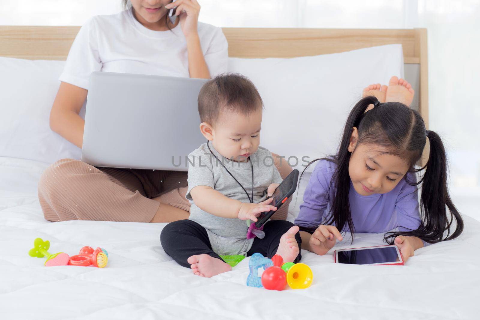 Young asian mother working at home online to internet with laptop computer and talking phone and daughter looking tablet on bed at bedroom, business woman is freelance, new normal, family concept.