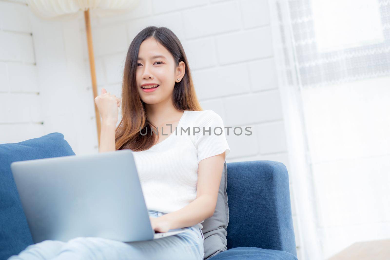 Young asian business woman smile and work from home with laptop computer online to internet on sofa in living room, freelance girl using notebook on couch with comfort, new normal, lifestyle concept.