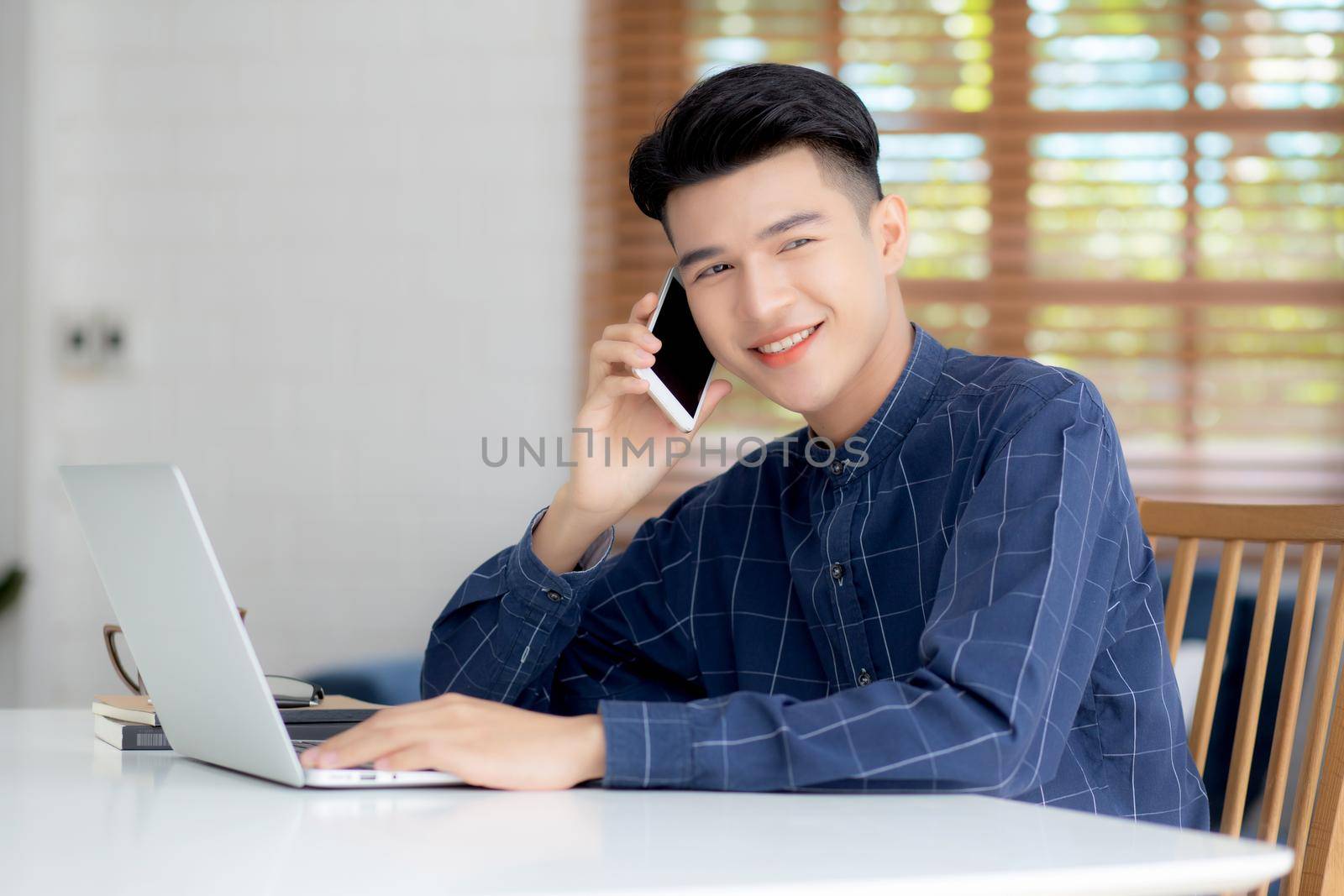 Young asian man talking phone and work from home with laptop computer, freelance using notebook to internet online and speak on smartphone, domestic life, business and communication concept. by nnudoo