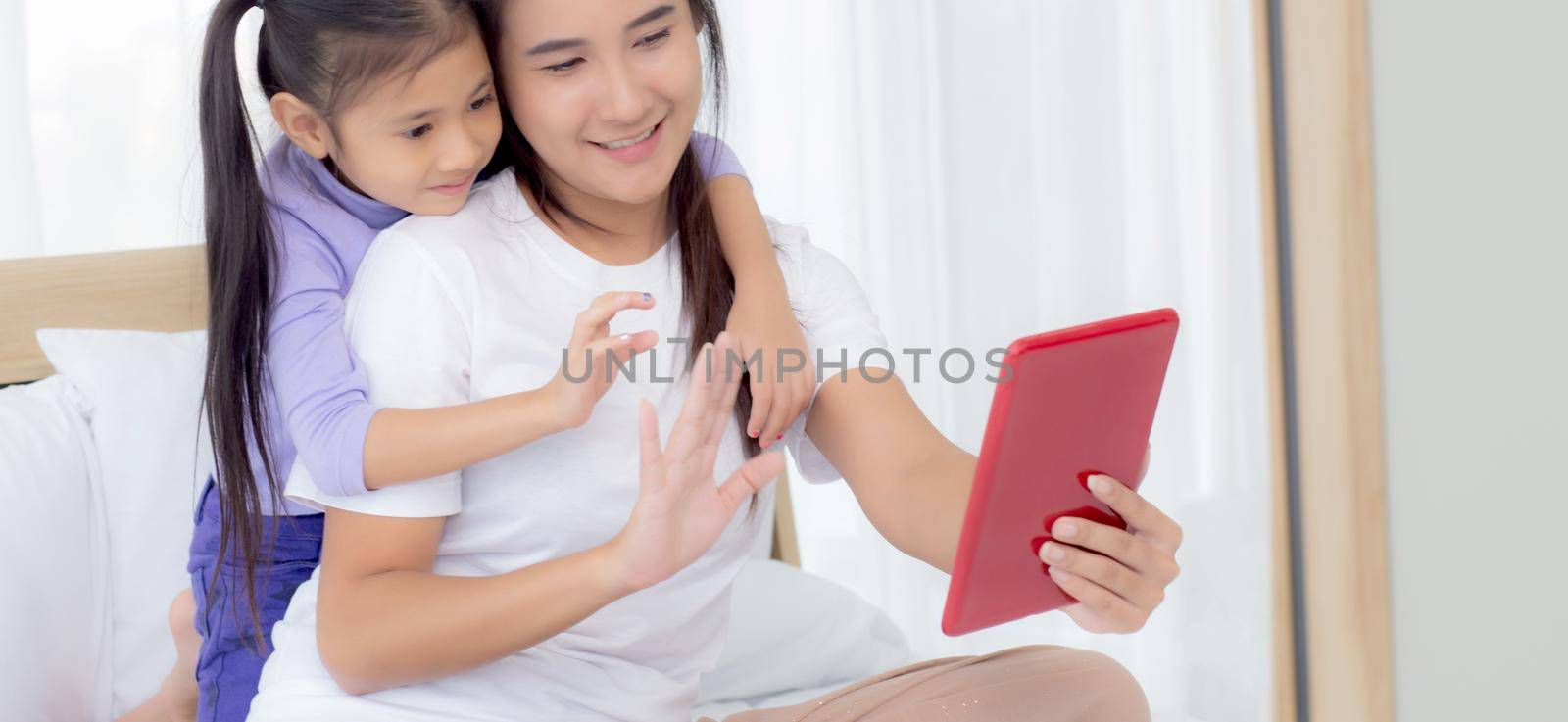 Young asian mom and daughter video call on tablet computer greeting with family together, happy mother and girl with relationship using technology in communication to internet online, two people. by nnudoo