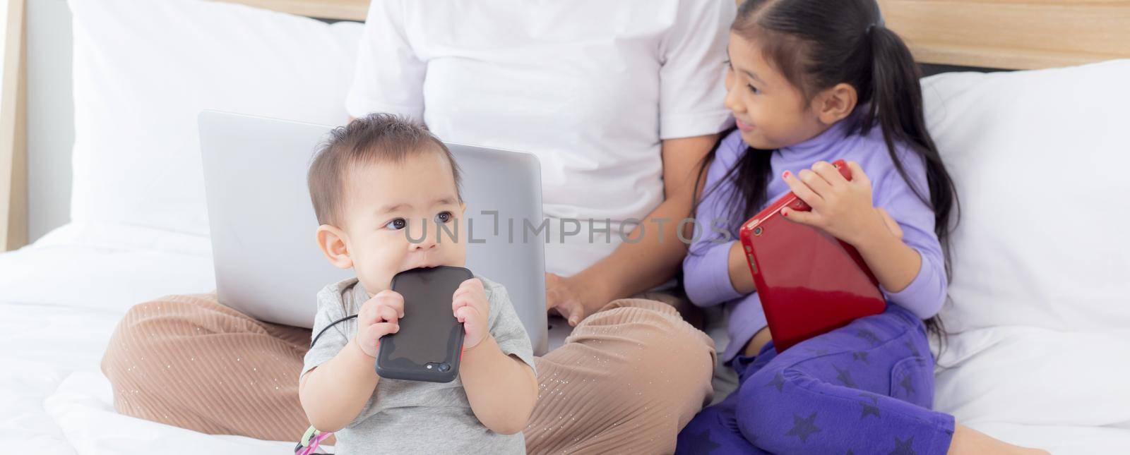 Young asian mother working at home online to internet with laptop computer and daughter looking tablet on bed at bedroom, business woman is freelance and children learning, new normal, family concept.