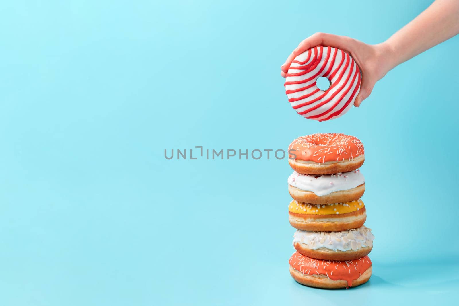 Stack of assorted donuts and one donut in female hand on blue background. Many colorful glazed doughnut with sprinkles stacked with copy space