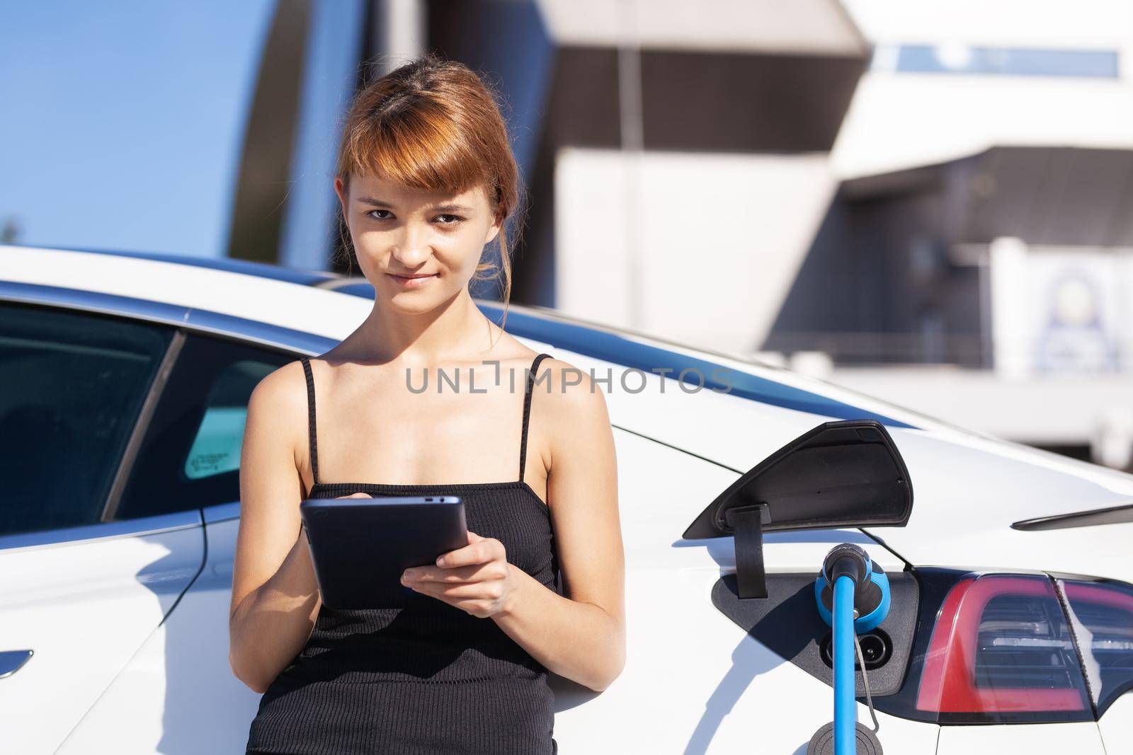 Girl working on tablet while her electric car is charging. by kokimk