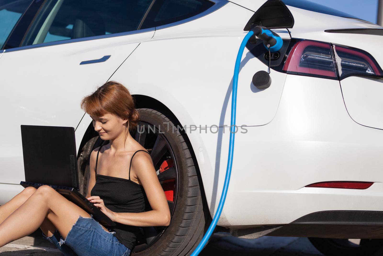 Girl waiting on the ground while her electric car is charging. Working on a laptop computer.