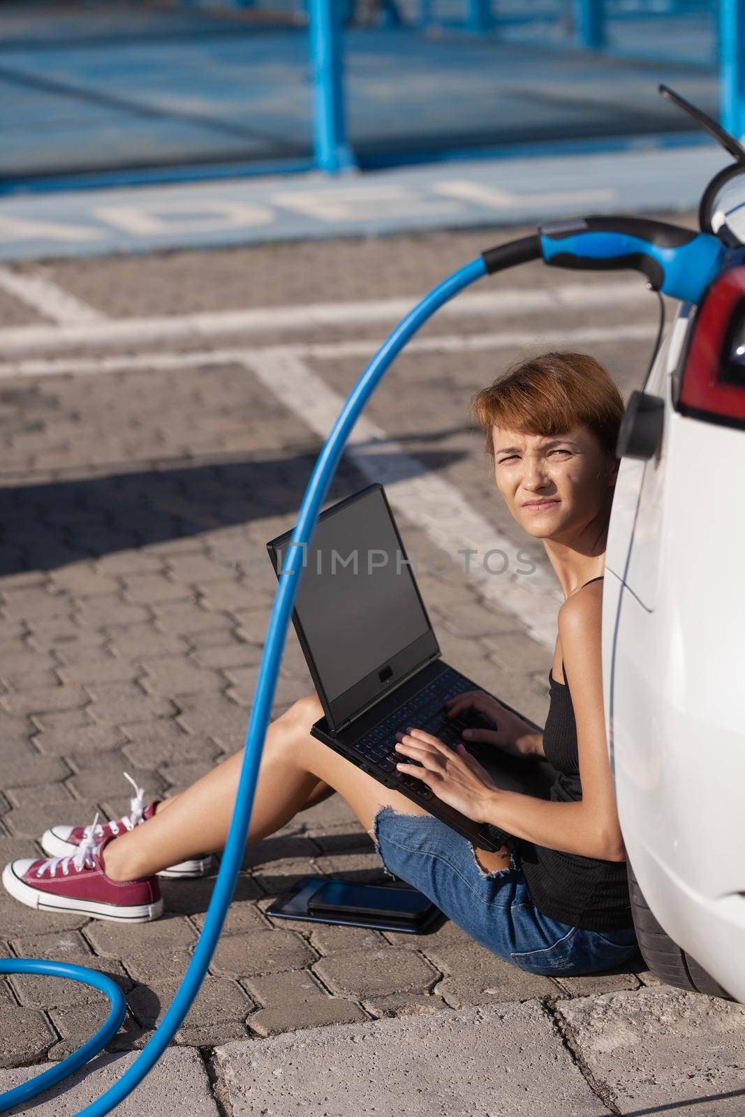 Girl waiting on the ground while her electric car is charging. Working on a laptop computer by kokimk