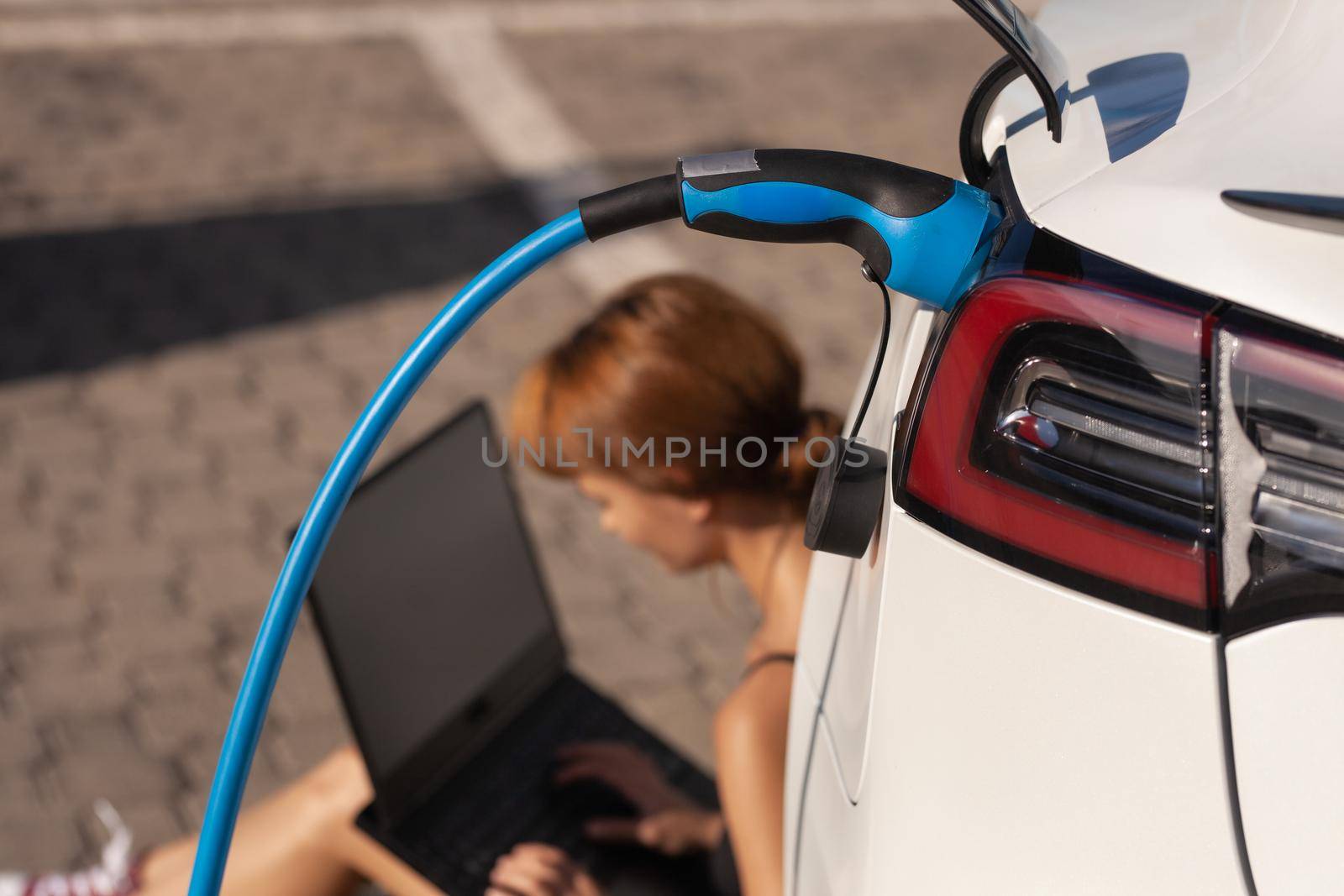 Girl waiting on the ground while her electric car is charging. Working on a laptop computer by kokimk