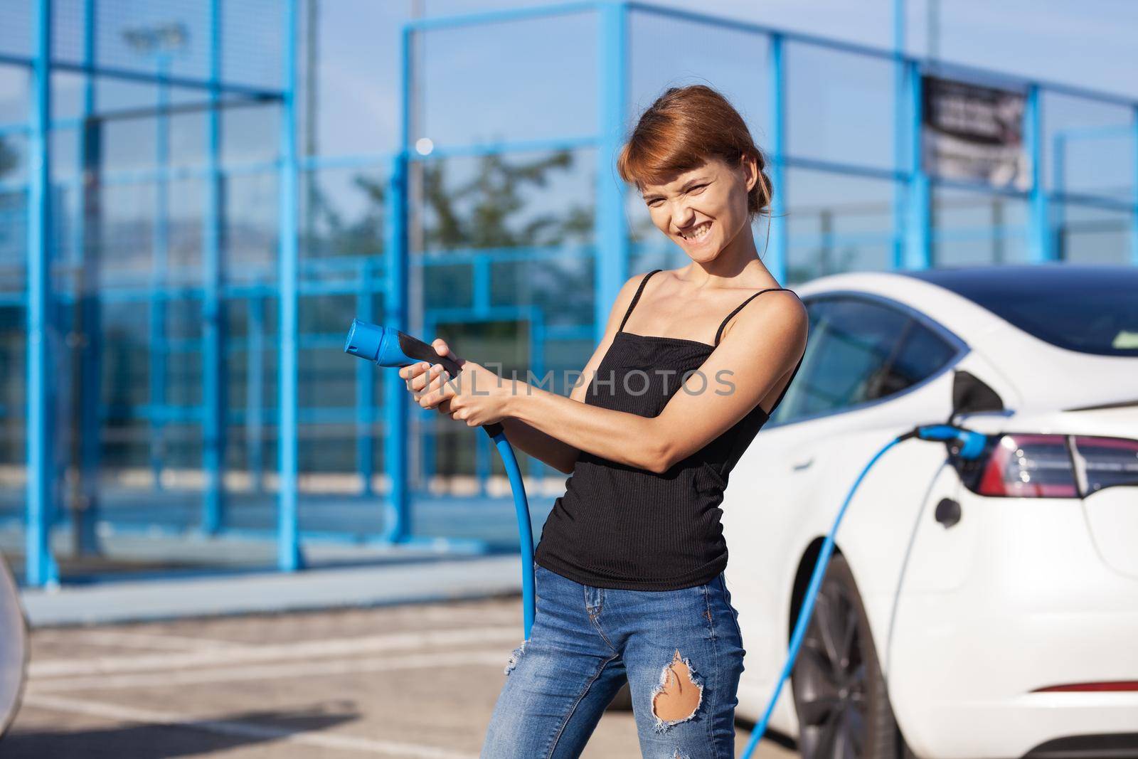 Beautiful young girl next to an electric car. Posing and holding a charging cable. by kokimk