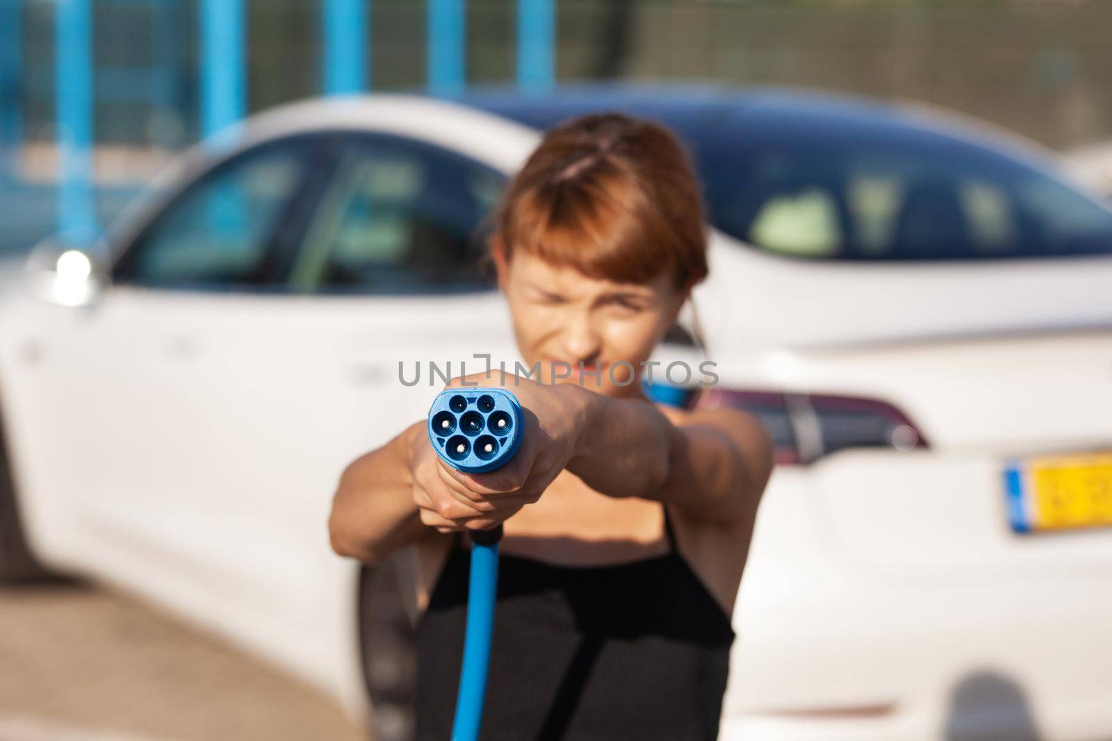 Beautiful young girl next to an electric car. Posing and holding a charging cable. by kokimk