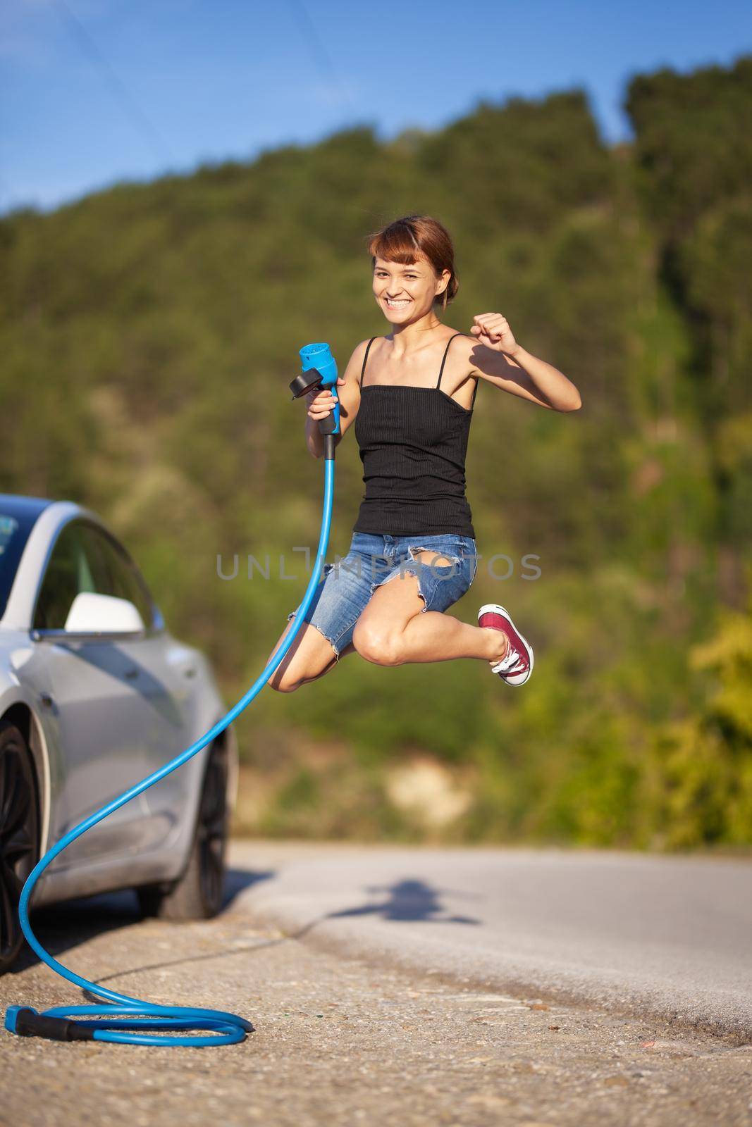 Beautiful young girl jumping next to an electric car. Holding blue charging cable by kokimk