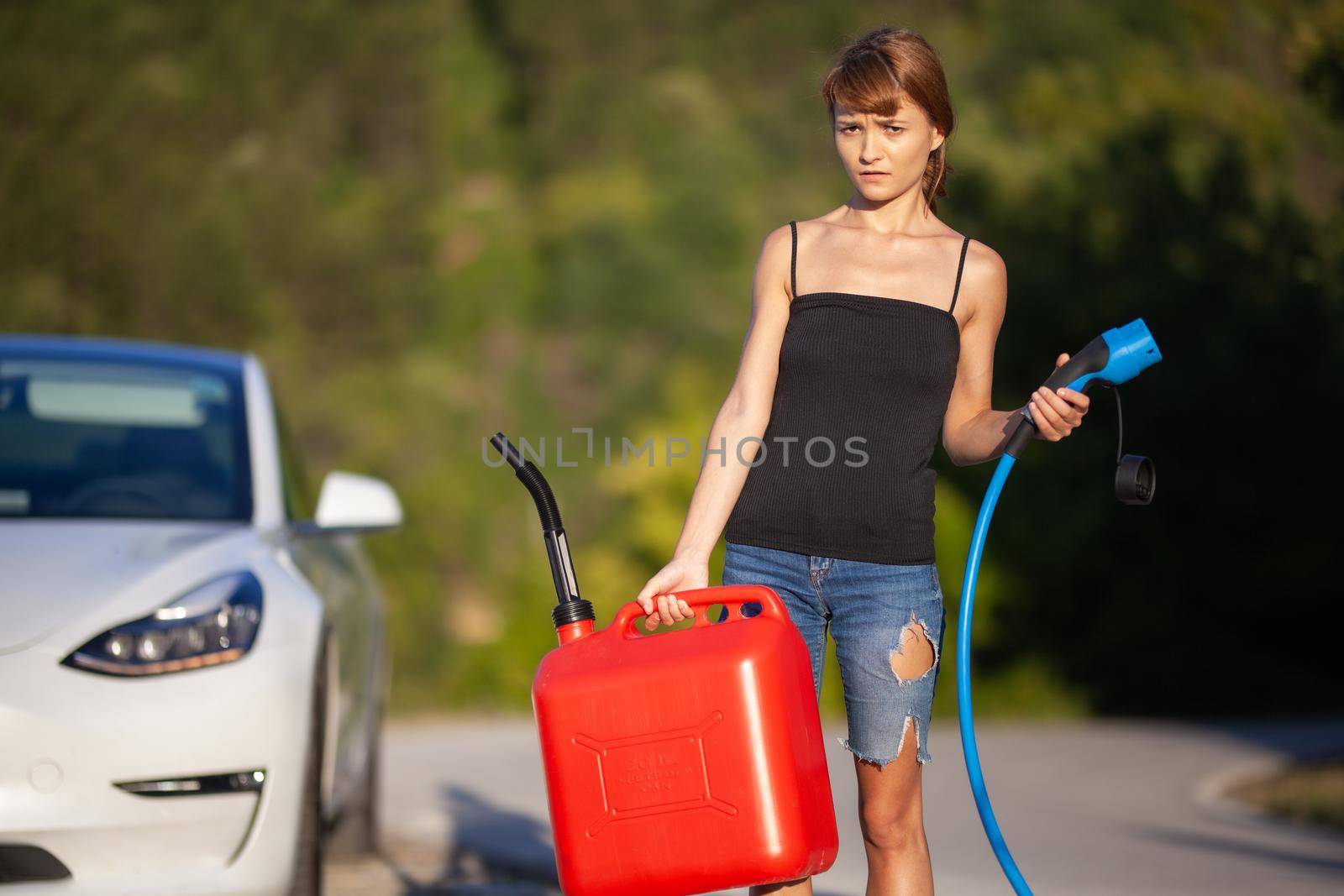 Confused girl standing next to an electric car. Holding charging cable and gassoline canister by kokimk