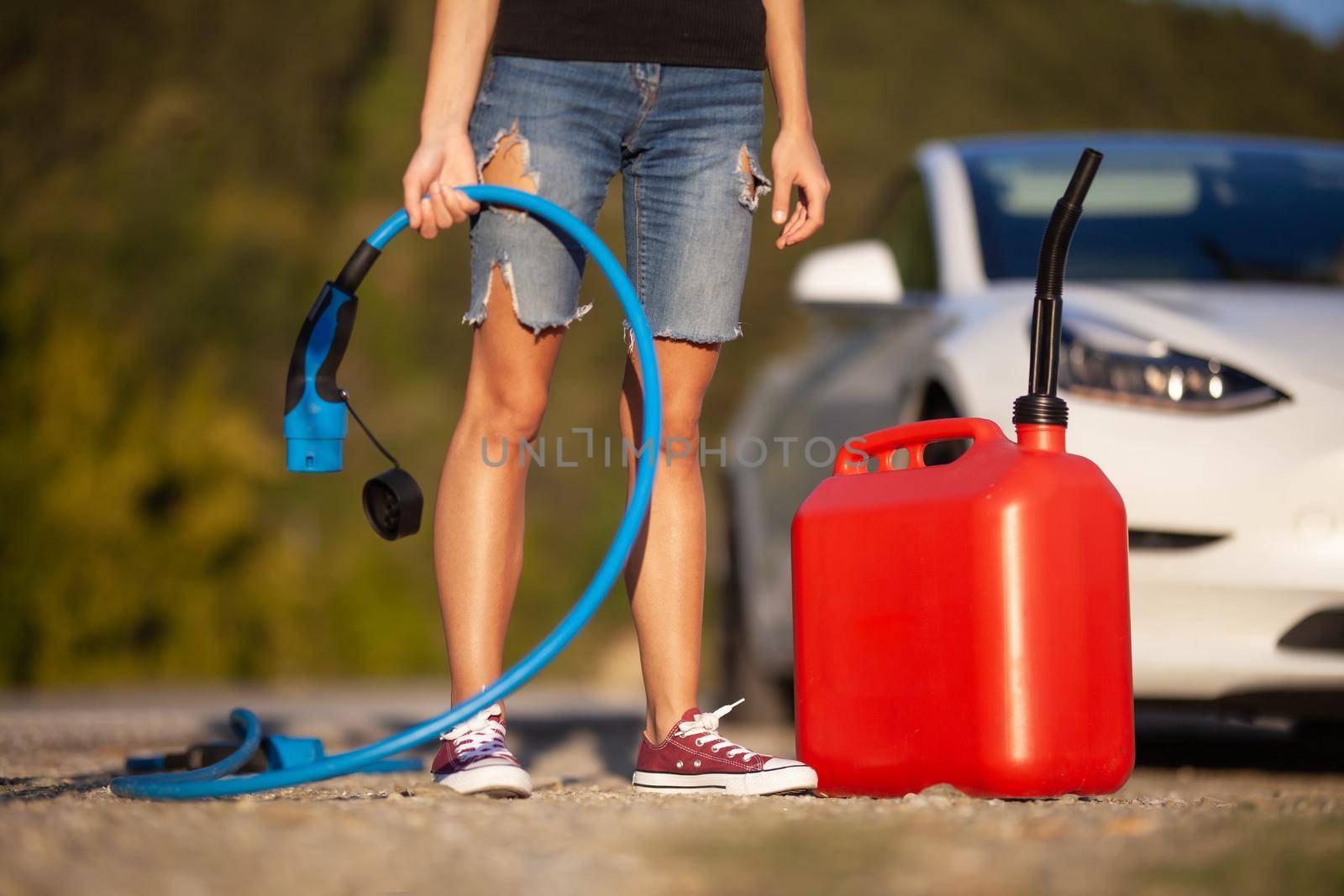 Girl standing next to an electric car. Holding charging cable and gassoline canister.