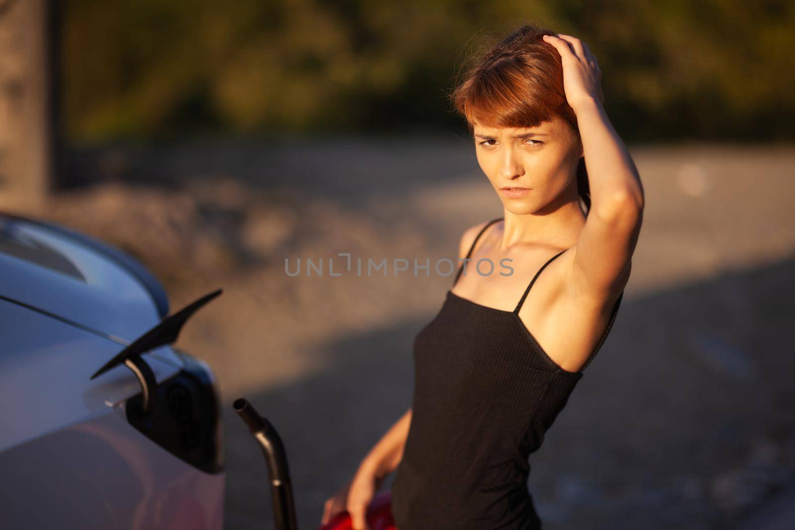 Confused girl trying to put gasoline from a red canister to an empty electric car charging socket by kokimk