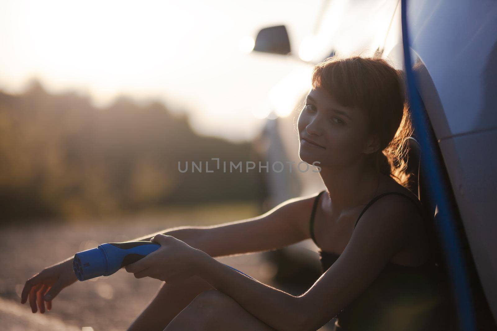 Beautiful young girl next to an electric car. Holding a charging cable. Sunset backlight by kokimk