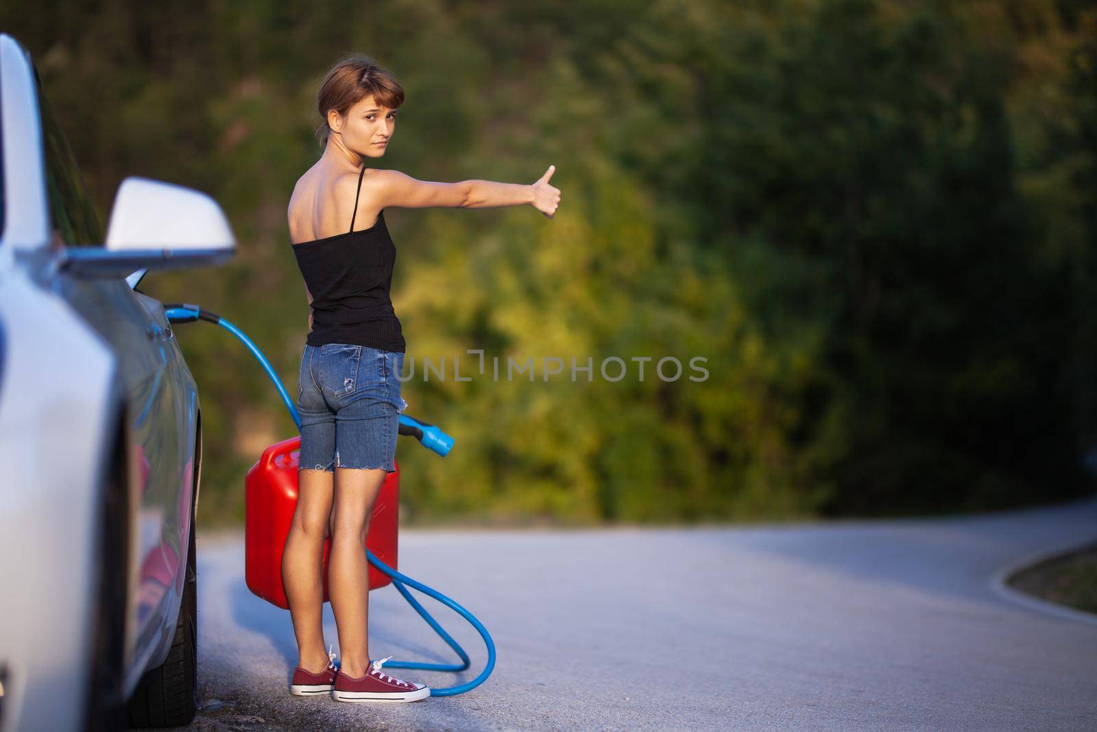 Girl standing next to electric car holding a charging cable and gassoline canister while hitchhiking.