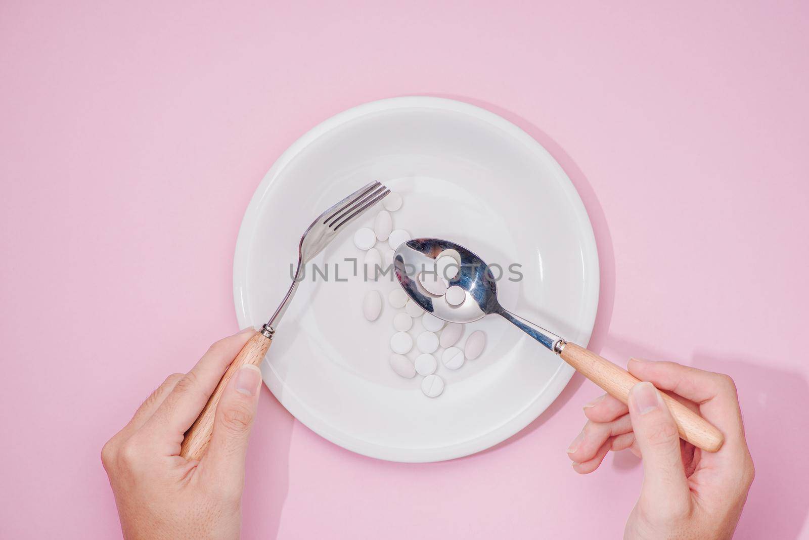 Top view of man's hands at dining table holding a fork and knife above dish with pills over pink background.