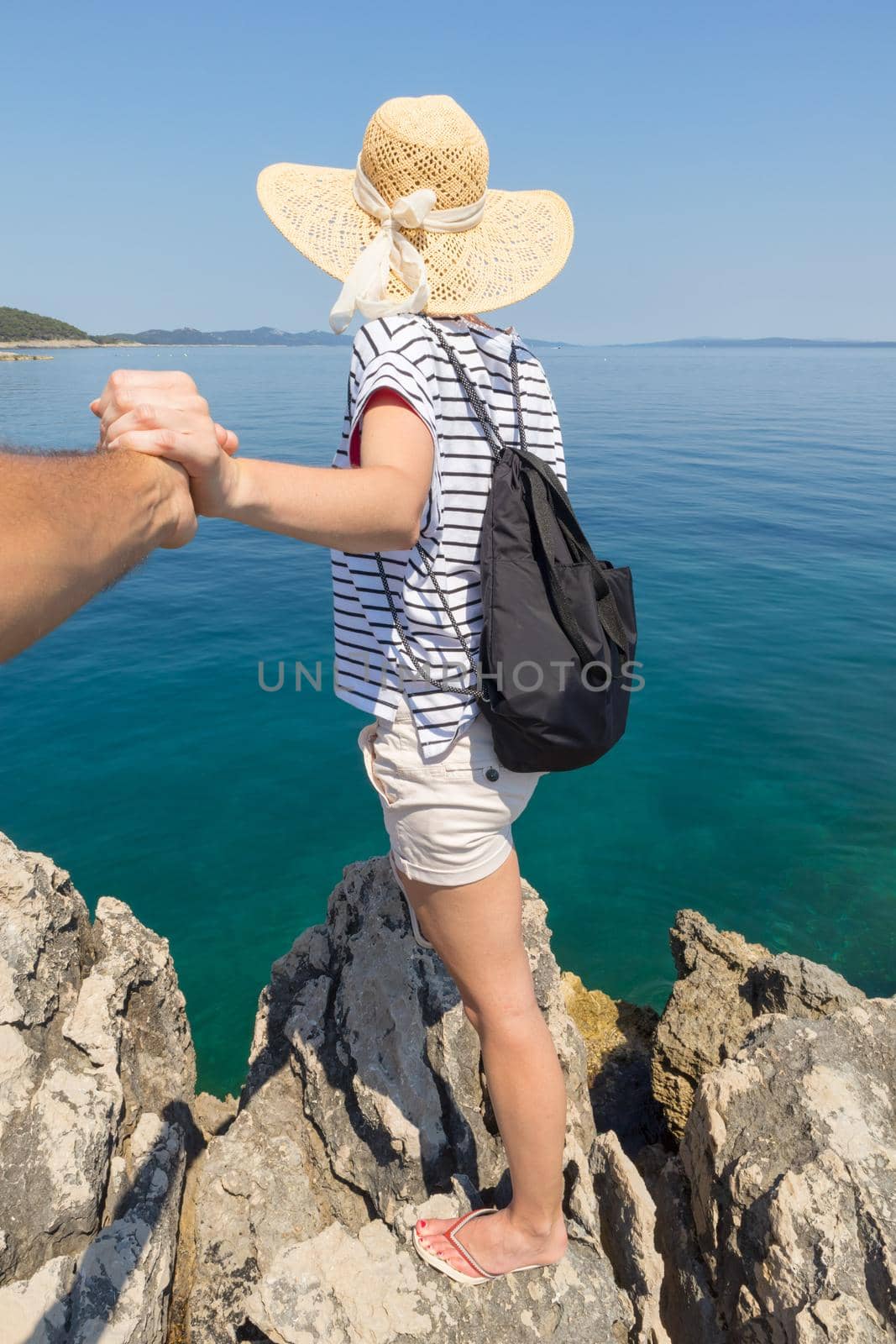 Woman traveler wearing straw summer hat and backpack, standing at edge of the rocky cliff, holding hand of her partner, looking at big blue sea and islands in on the horizon