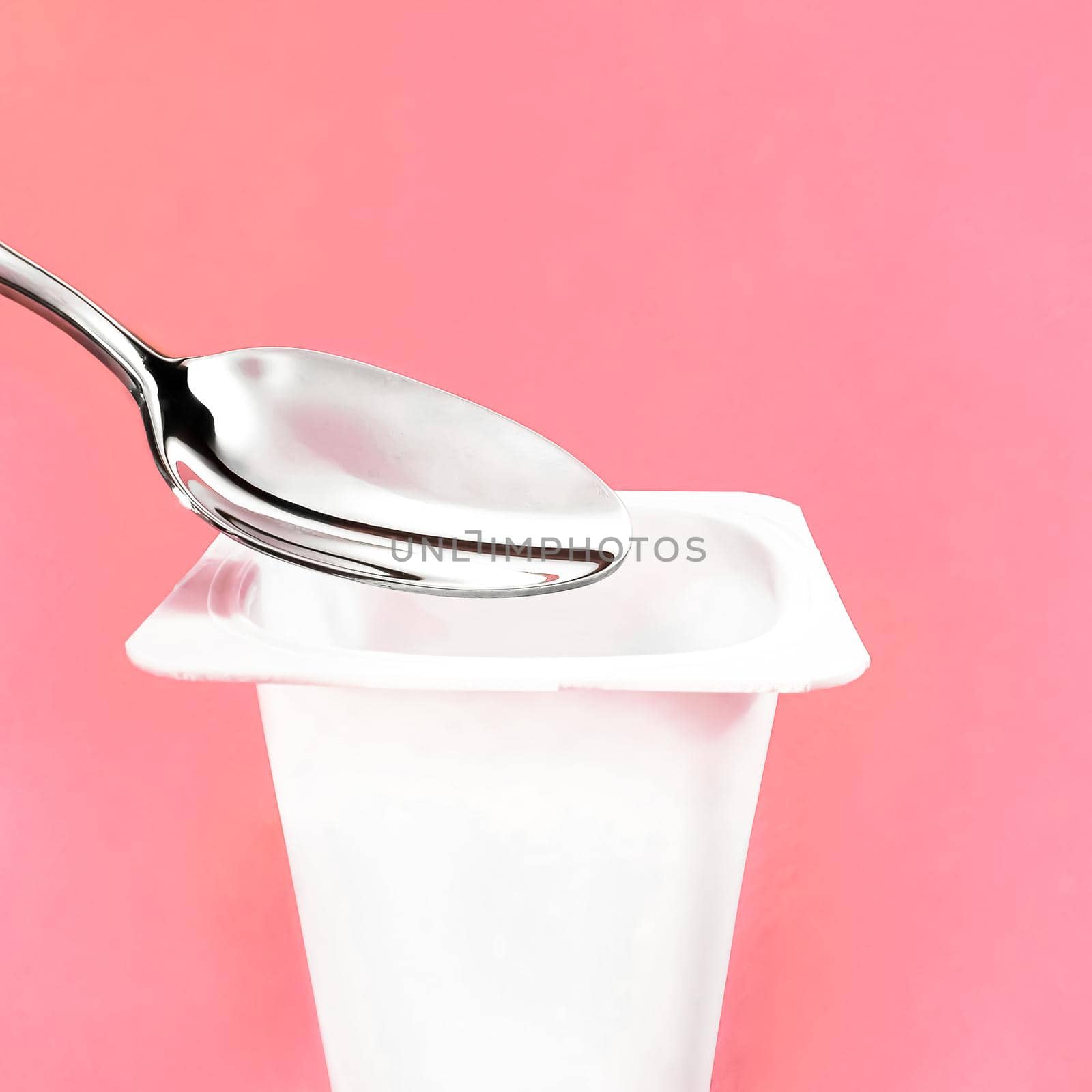 Yogurt cup and silver spoon on pink background, white plastic container with yoghurt cream, fresh dairy product for healthy diet and nutrition balance.