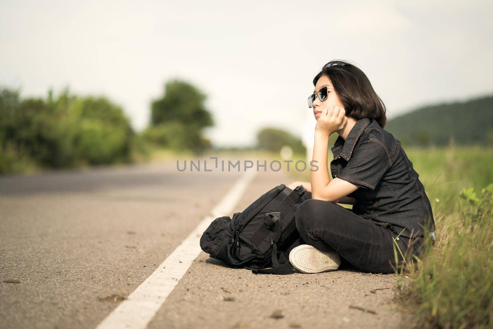 Woman sit with backpack hitchhiking along a road in countryside by stoonn