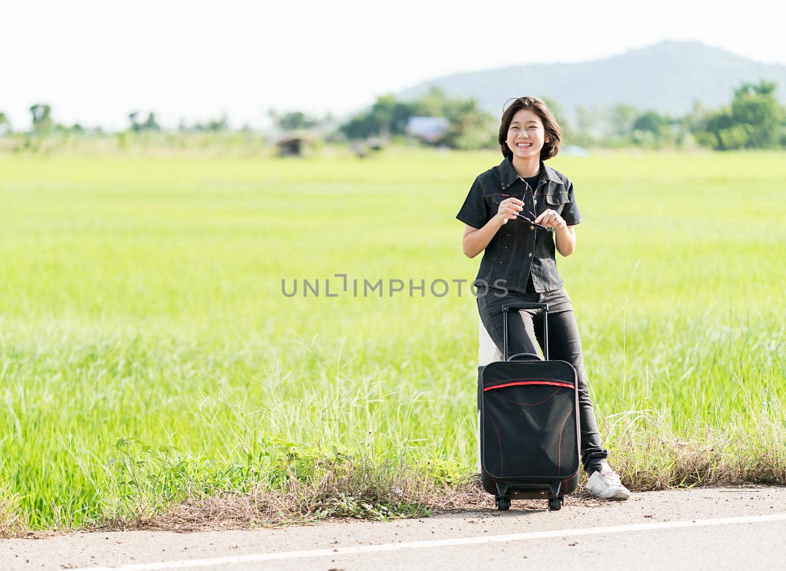 Young asian woman short hair and wearing sunglasses with luggage hitchhiking along a road in countryside Thailand
