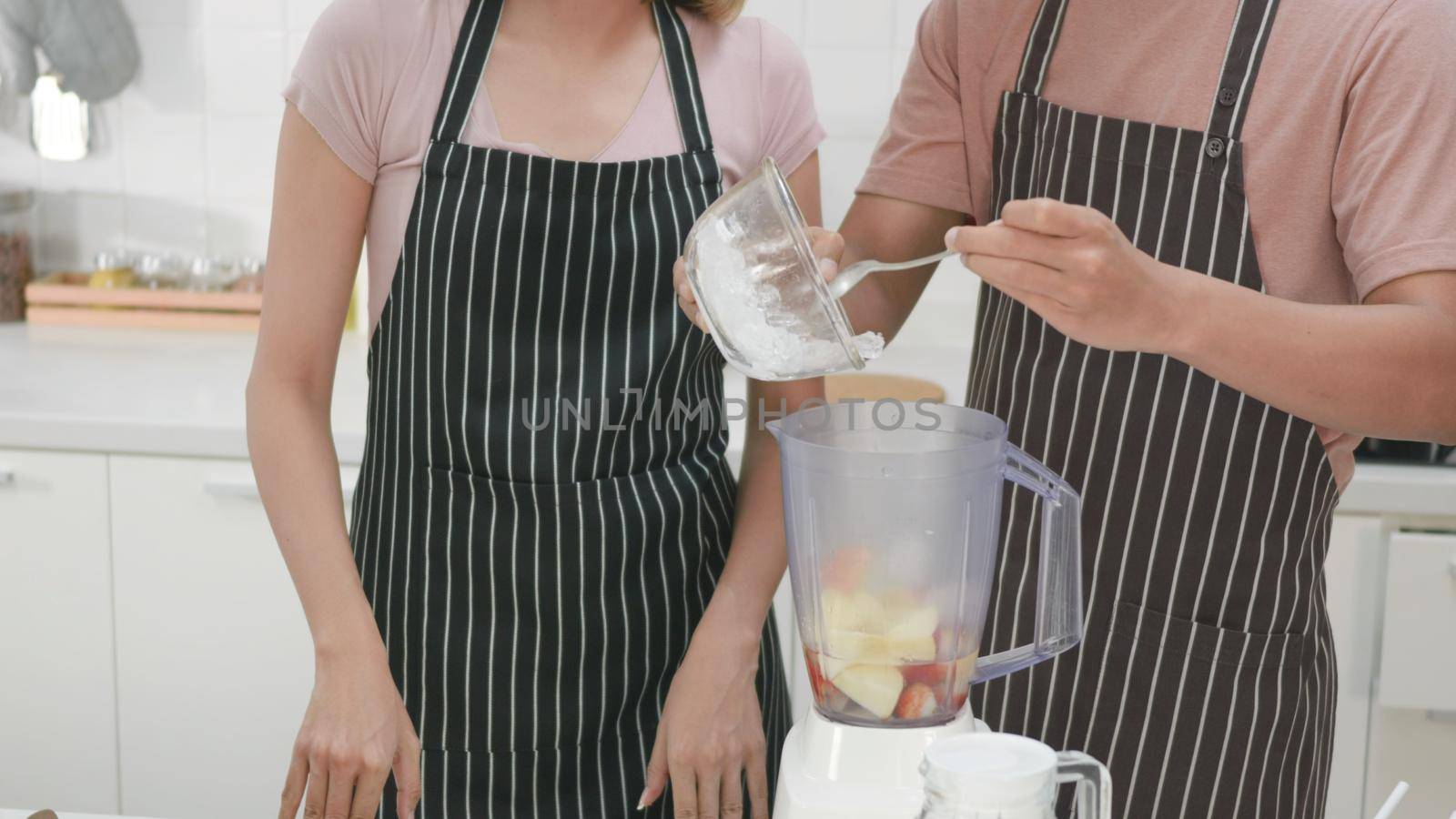 Happy Asian beautiful young family couple husband and wife making fresh apple smoothie in kitchen together at home. The man and woman pour ice into a juice maker blender. Healthy lifestyle concept
