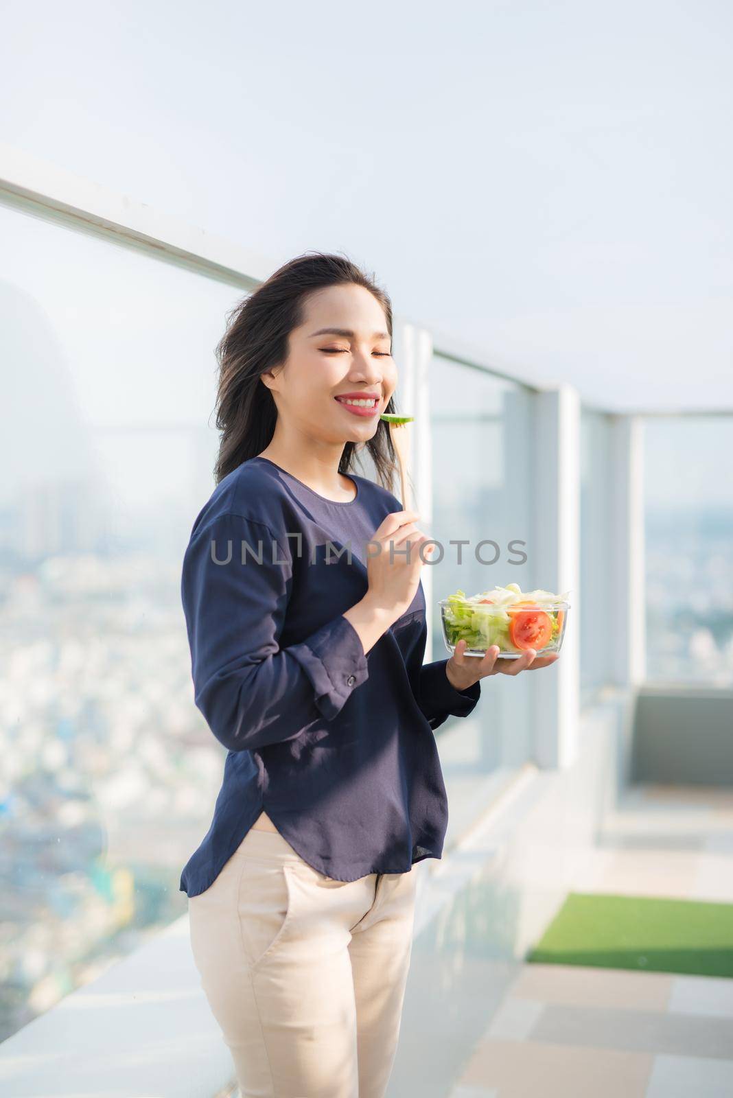 Portrait of attractive caucasian smiling woman eating salad on a sunny summer day sitting on green grass in park, focus on fork with salad by makidotvn