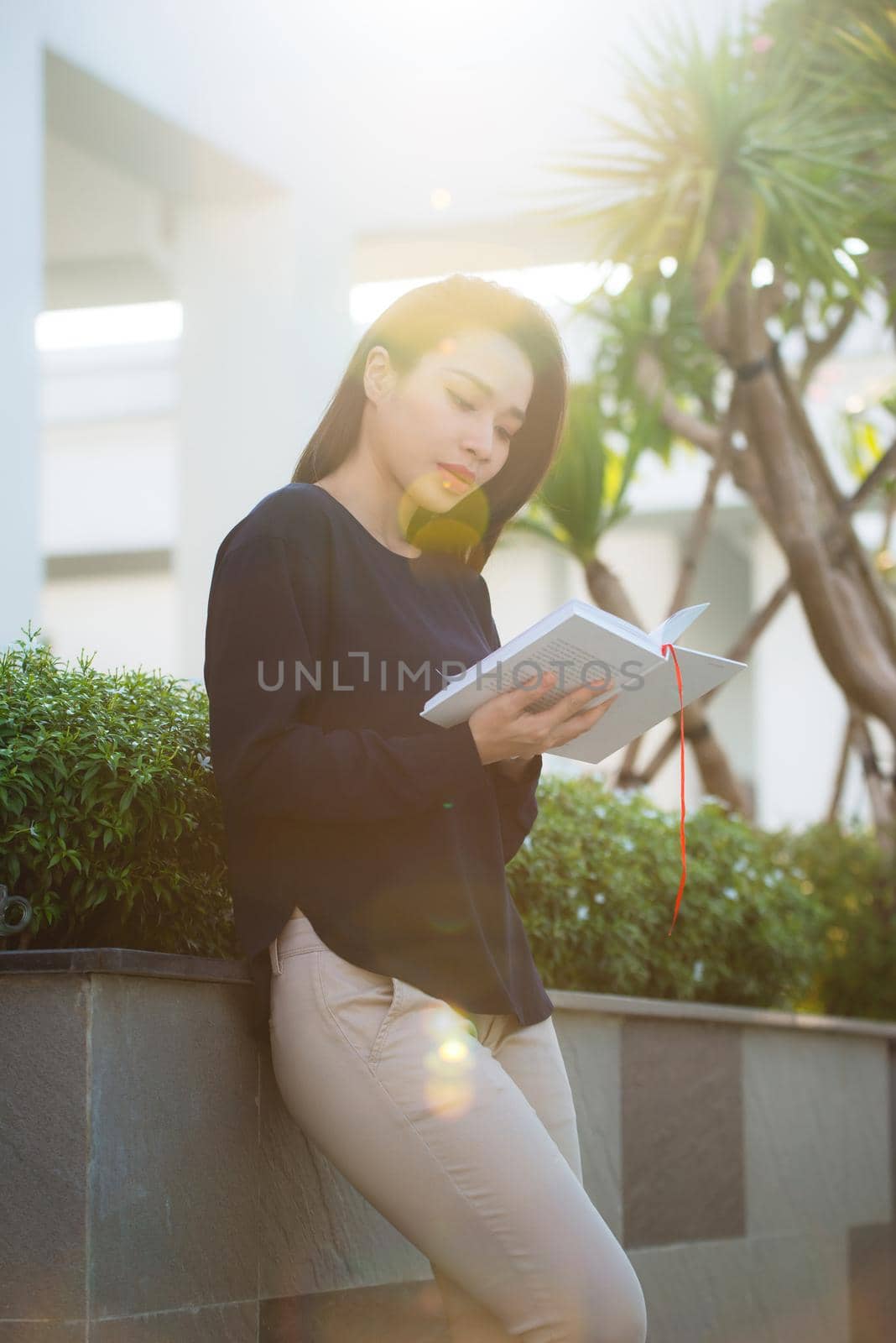 Portrait of smiling female student reading book while standing outdoor on terrace of campus cafe in sunny day. Education, lifestyle and people concept.