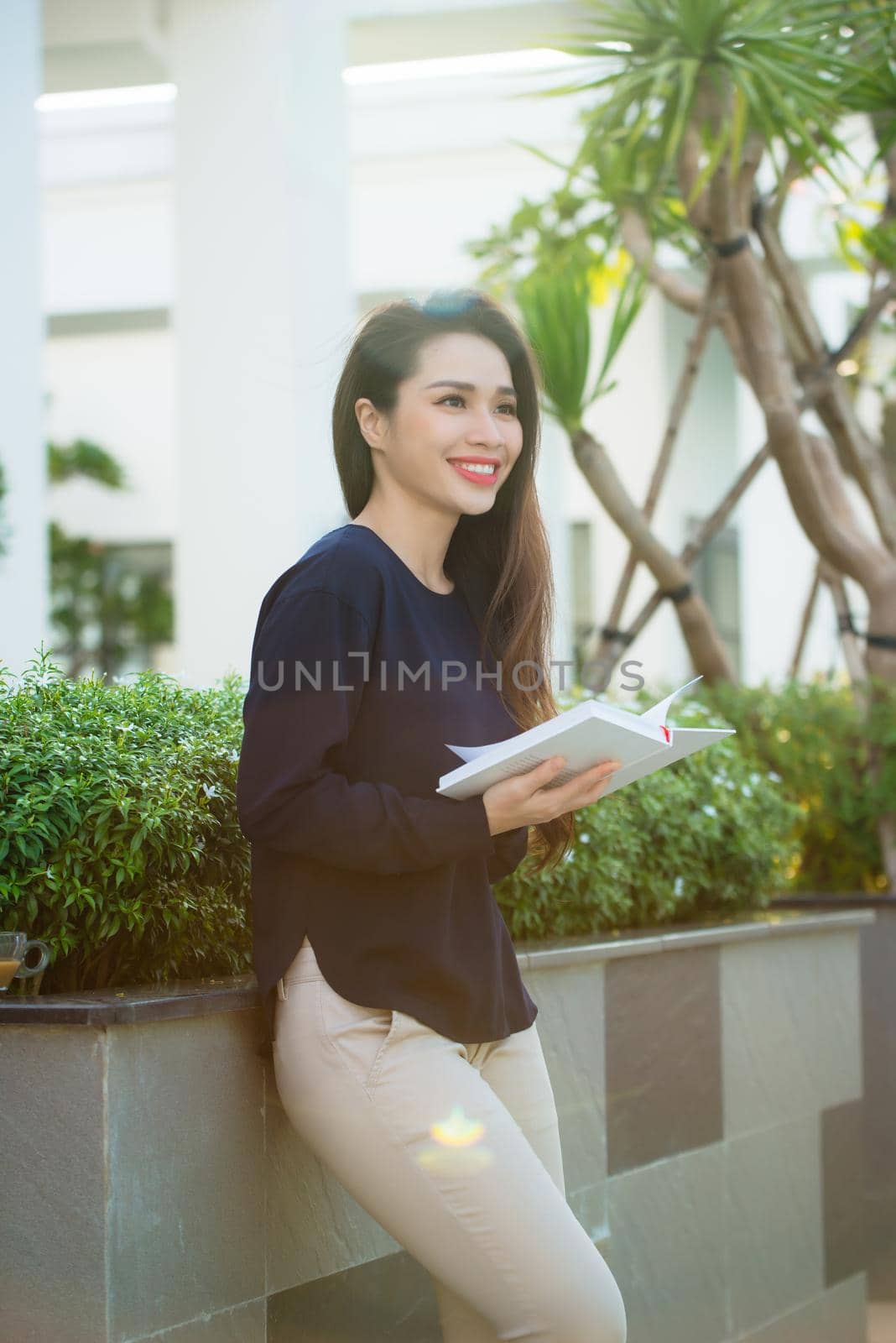 Portrait of smiling female student reading book while standing outdoor on terrace of campus cafe in sunny day. Education, lifestyle and people concept.