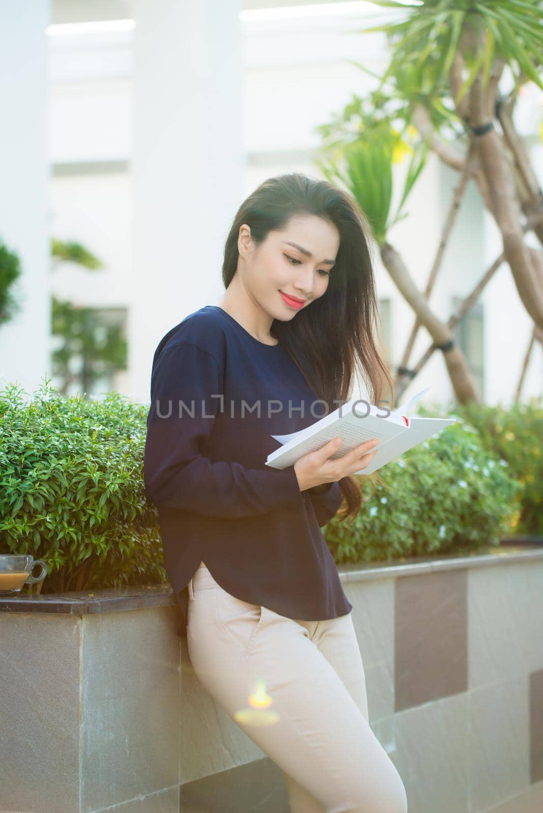 Portrait of smiling female student reading book while standing outdoor on terrace of campus cafe in sunny day. Education, lifestyle and people concept.