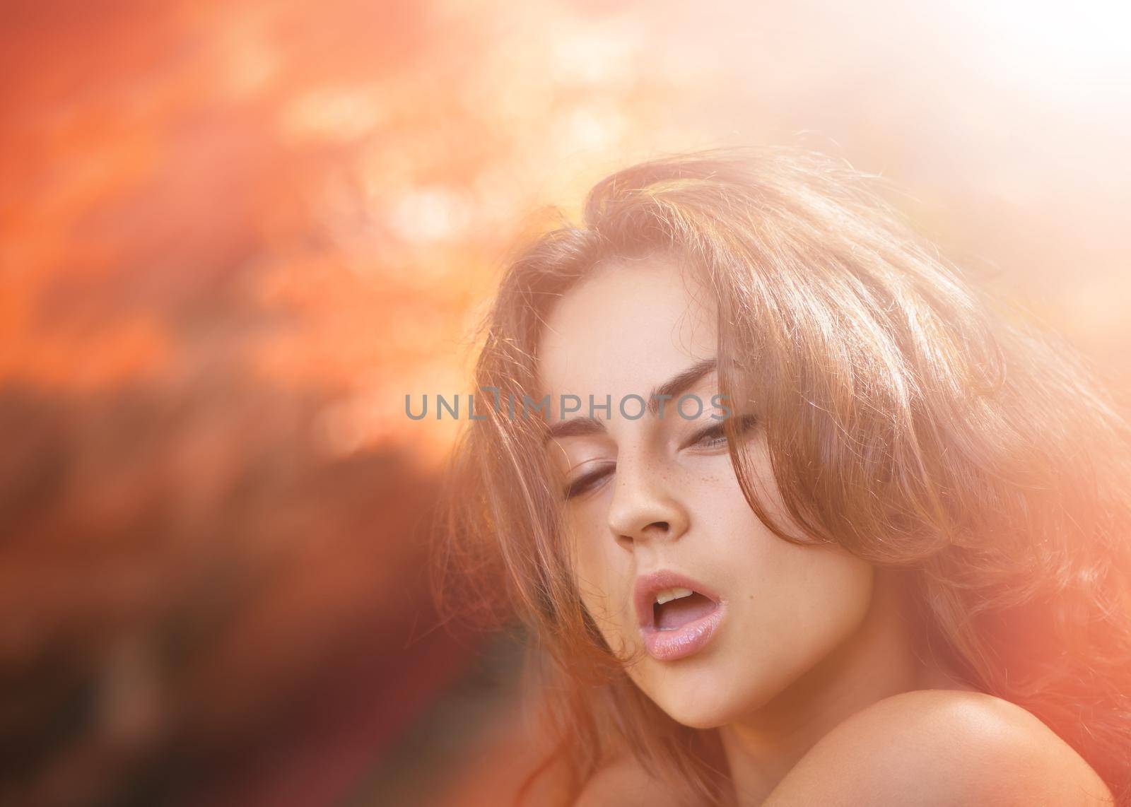 Portrait of an emotional young woman in sun light against red blured nature background of autumn forest.