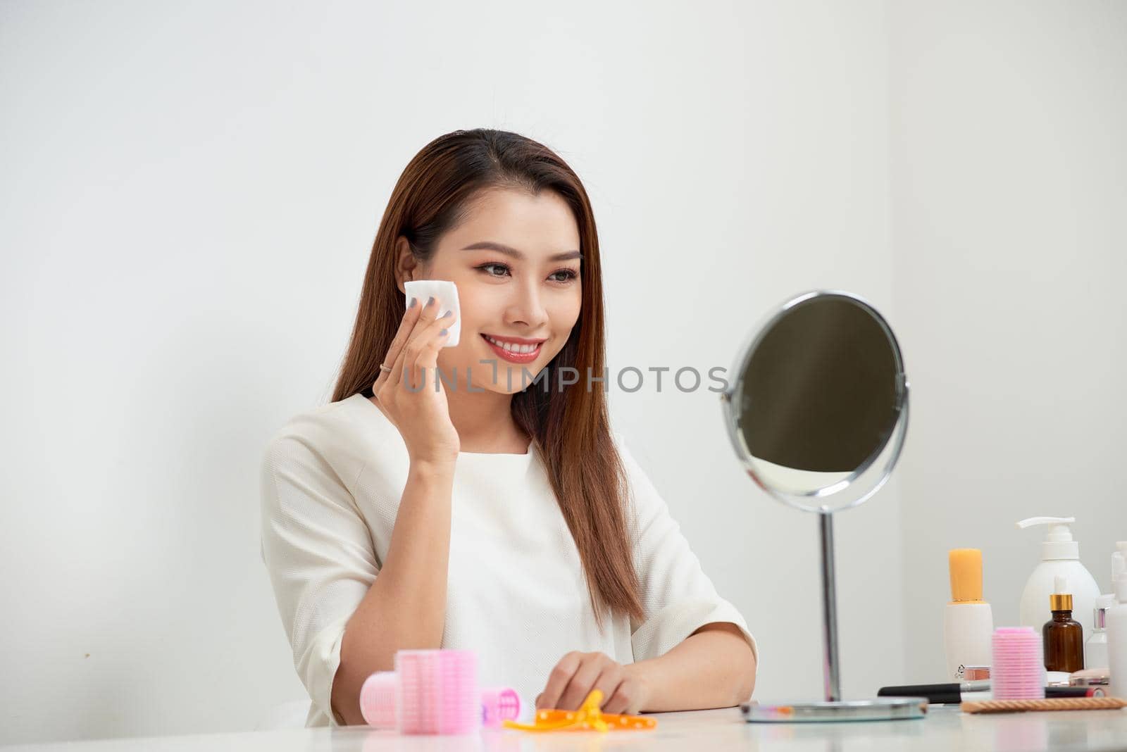 Taking off her make-up. Beautiful cheerful young woman using cotton disk and looking at her reflection in mirror with smile while sitting at the dressing table