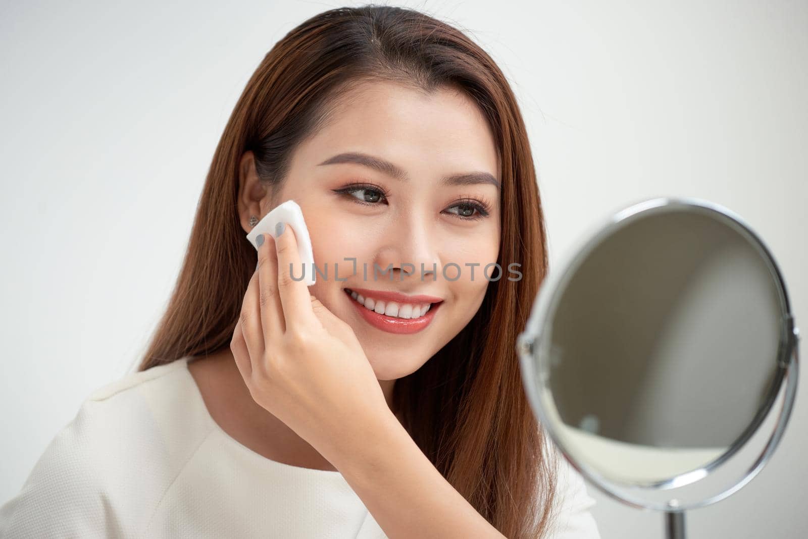 Taking off her make-up. Beautiful cheerful young woman using cotton disk and looking at her reflection in mirror with smile while sitting at the dressing table by makidotvn