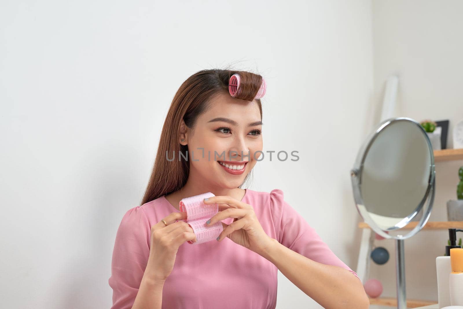 Head and shoulders portrait of beautiful Asian woman wearing hair curlers looking in mirror with wide smile, home interior on background